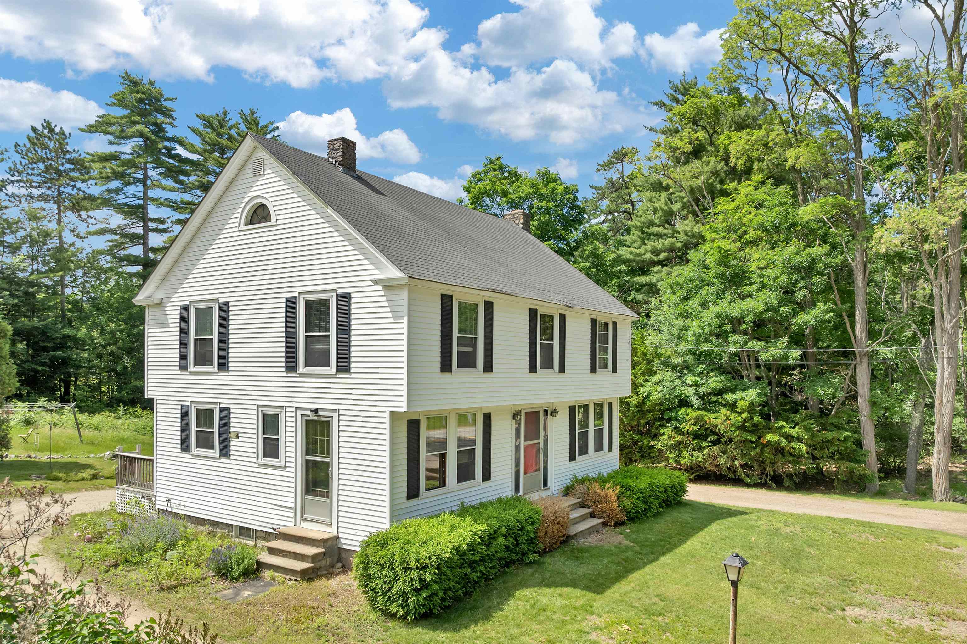 White two-story house with black shutters, surrounded by mature trees and a green lawn.