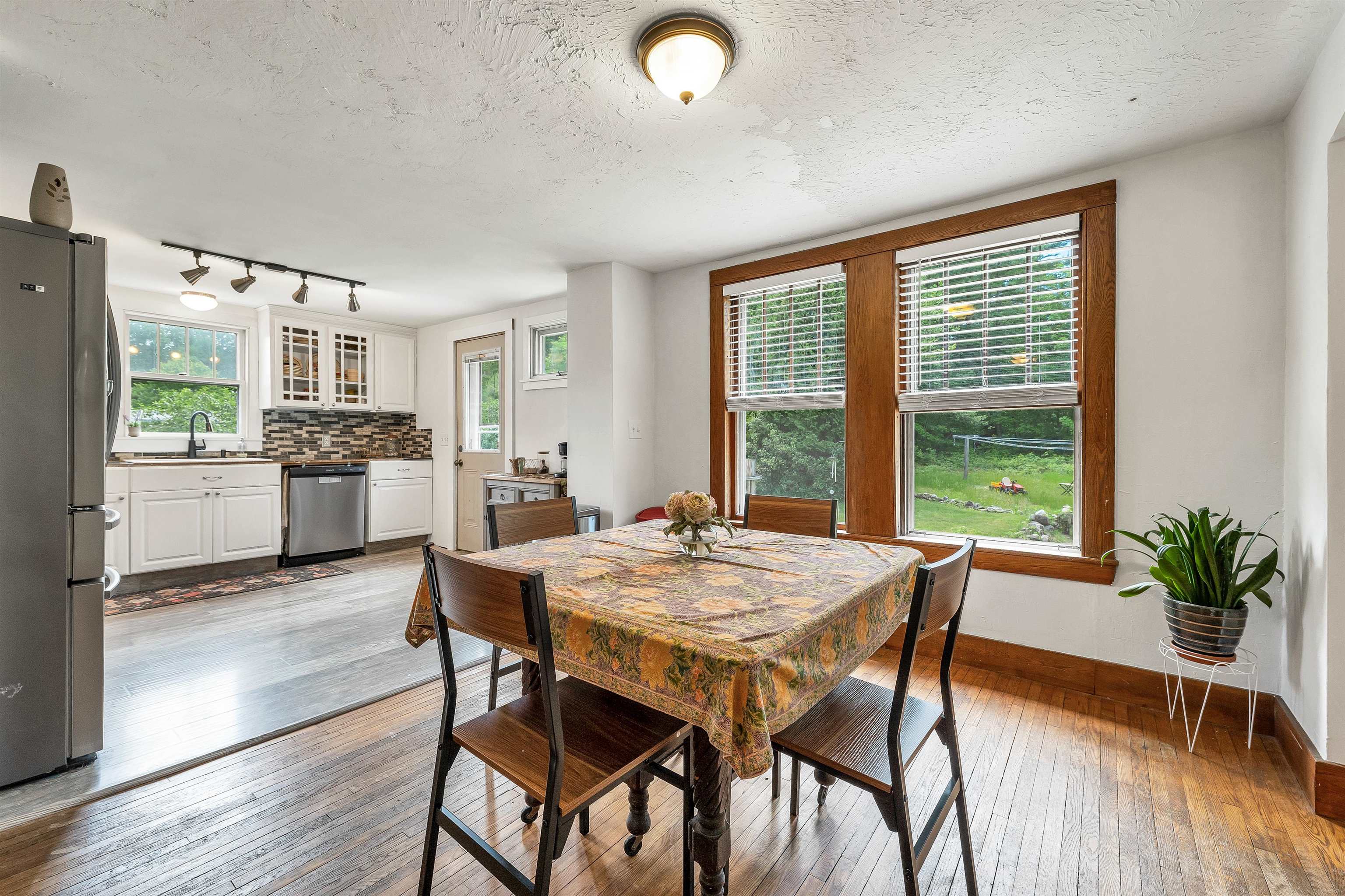 Open kitchen and dining area with white cabinets, hardwood floors, and large wood-trimmed windows overlooking a yard.