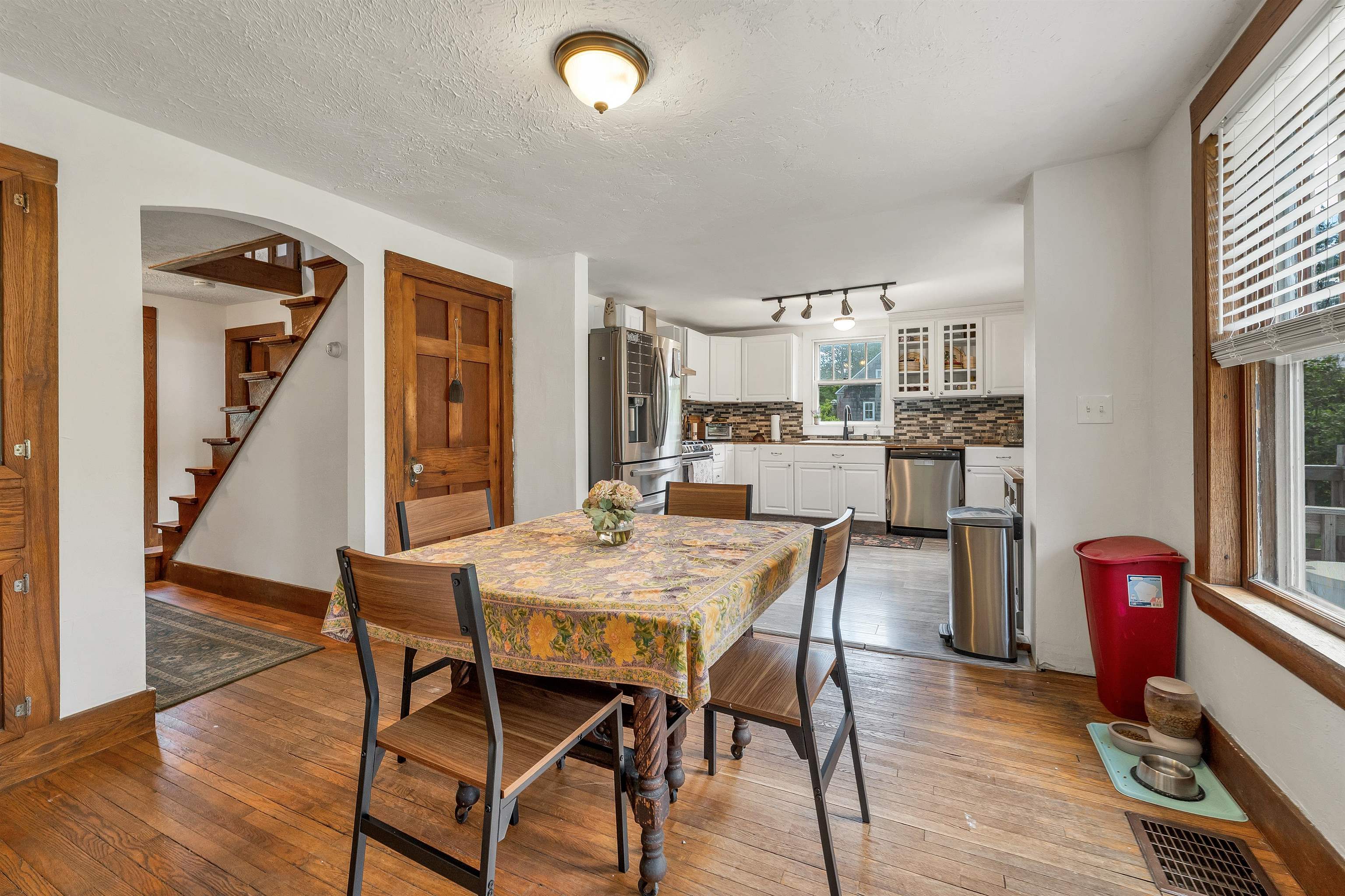 Dining area with a floral tablecloth, hardwood floors, and an open kitchen with white cabinets and stainless appliances.