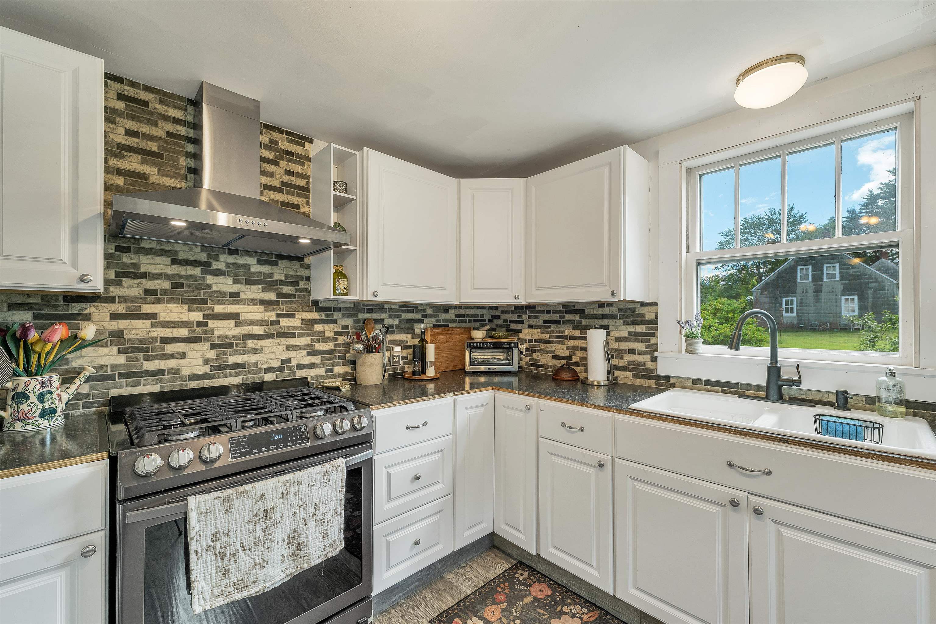 Kitchen with white cabinets, a brick backsplash, gas range, and farmhouse sink overlooking a green yard.