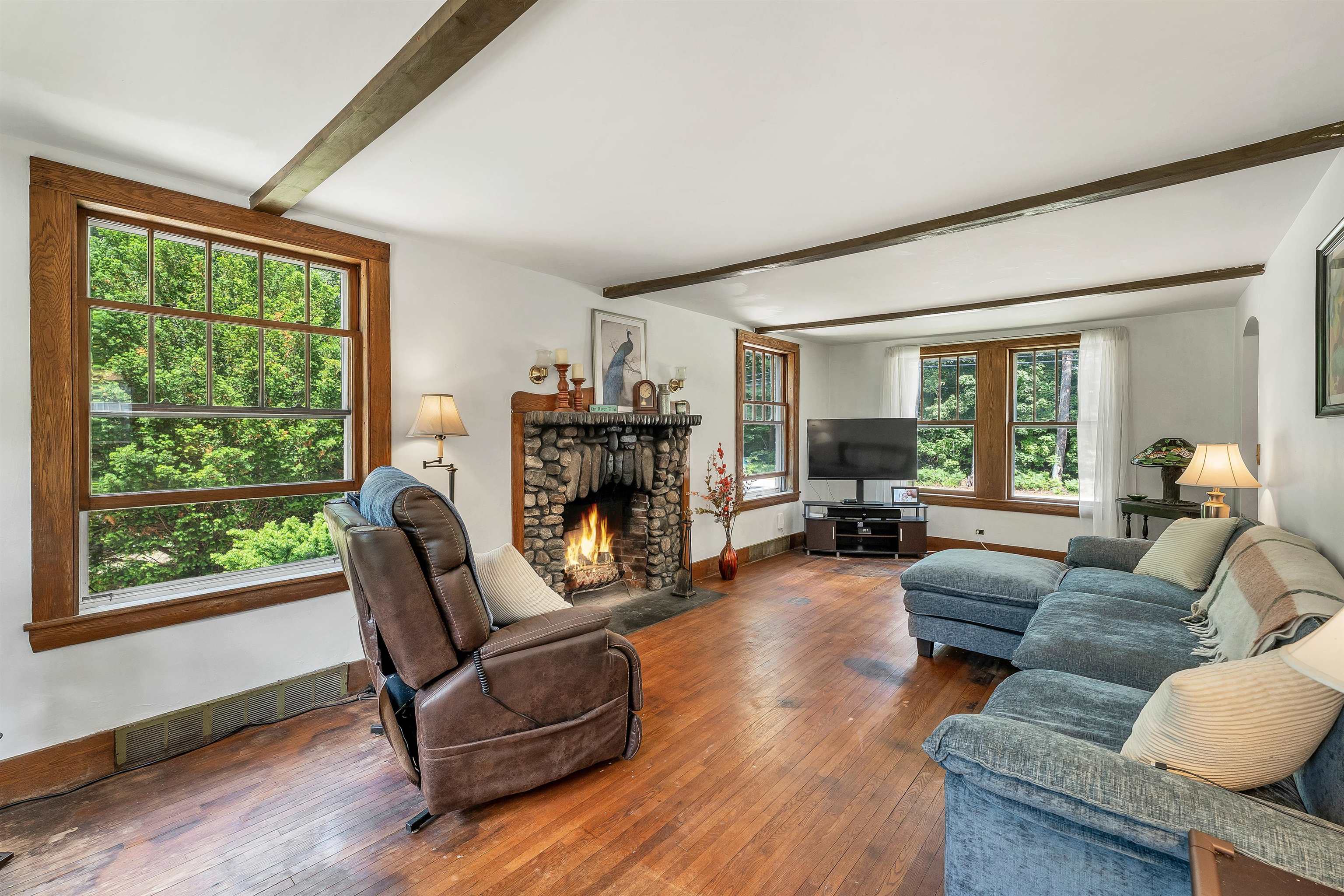 Living room with a stone fireplace, exposed beams, and large windows overlooking green trees.