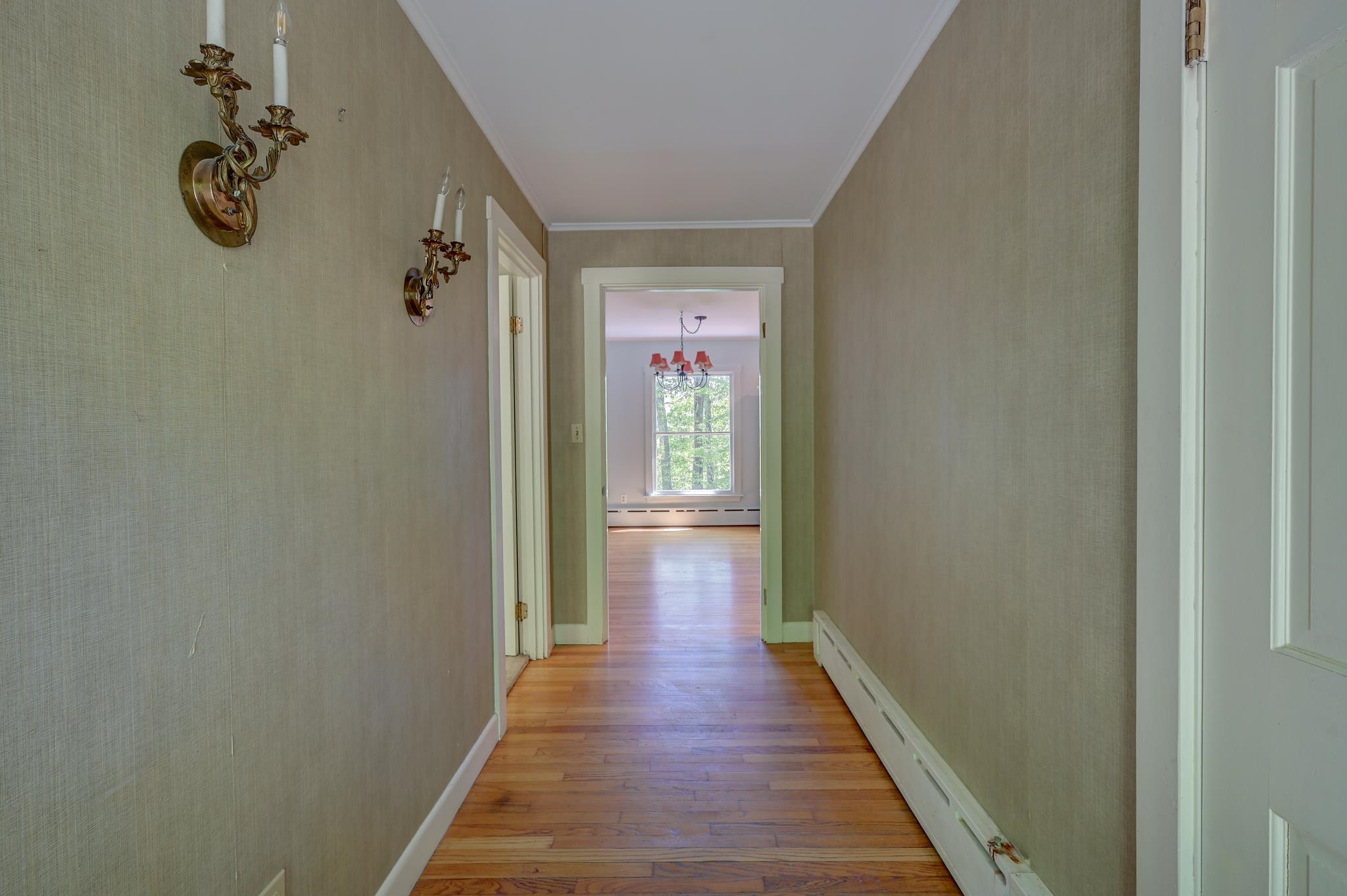Hallway with hardwood floors and brass sconces leading to a bright room with a chandelier and window.