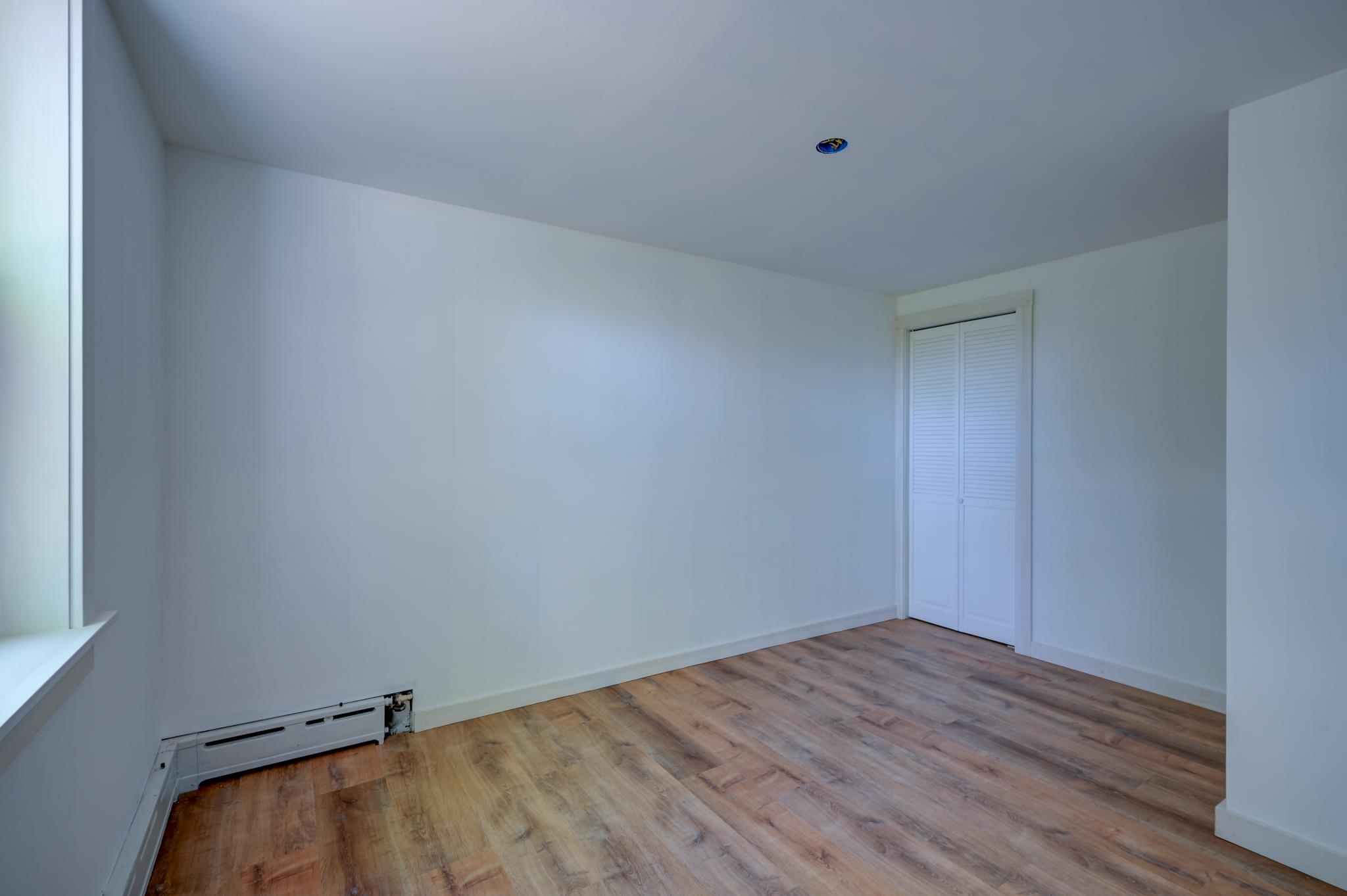 Empty bedroom with wood-look flooring, a window, and a louvered-door closet.