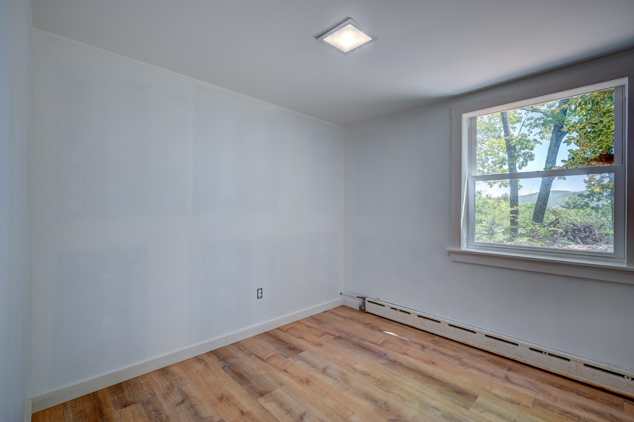 Empty room with wood-look flooring, a long baseboard heater, and a window overlooking trees and a wooded hill.