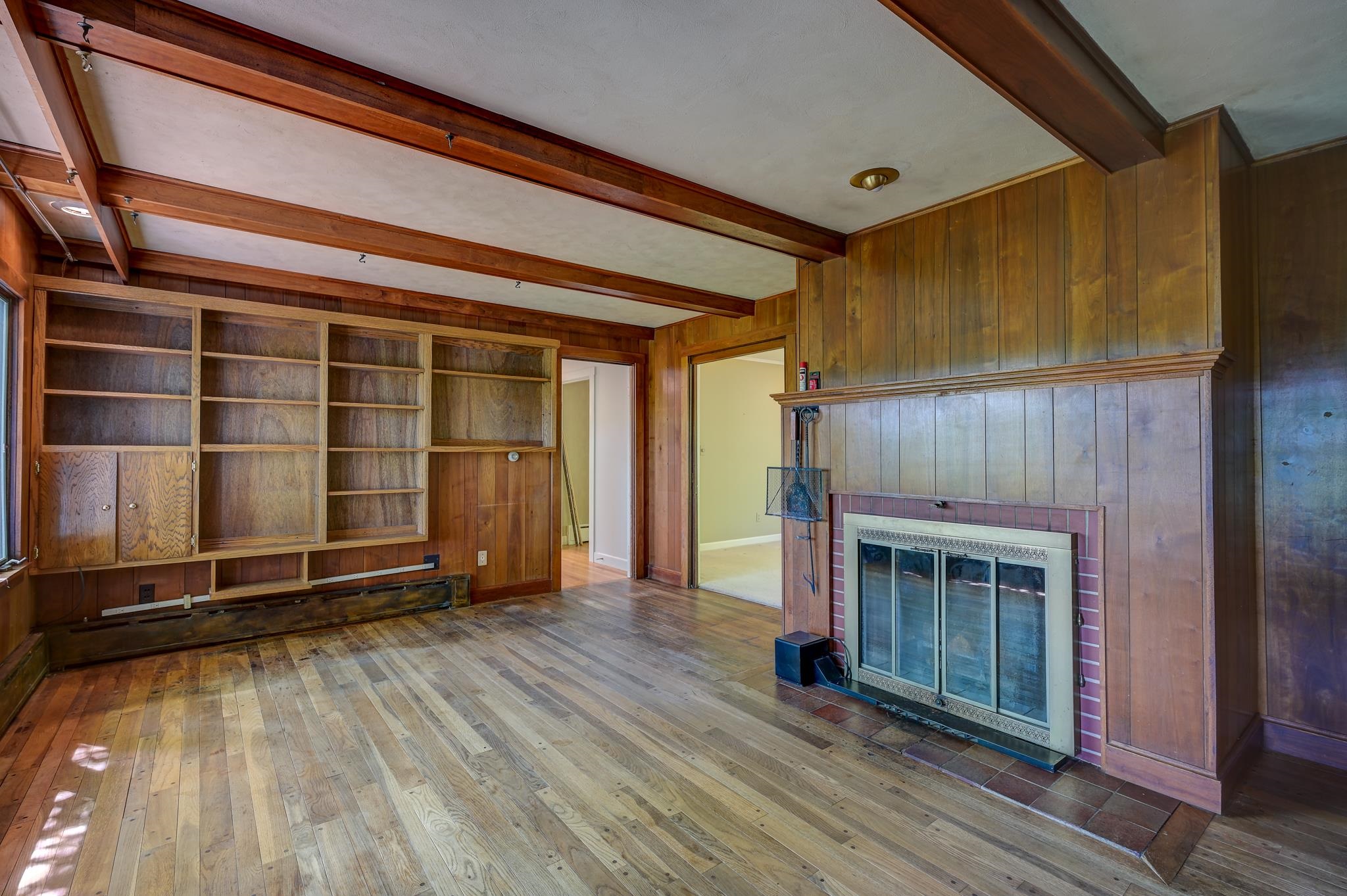 Empty wood-paneled room with built-in shelving, exposed beams, and a tiled fireplace.