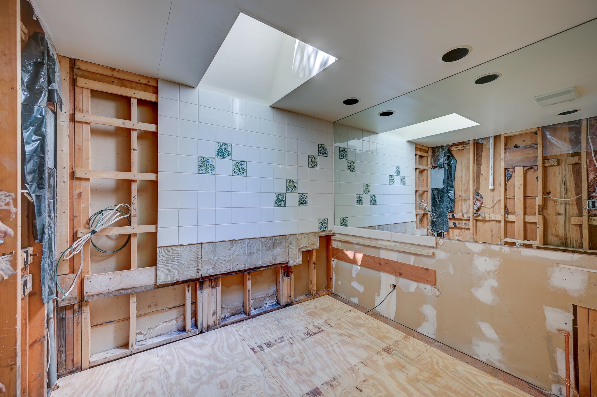 Bathroom under renovation with exposed framing, floral accent tiles, a large mirror, and ceiling skylights.
