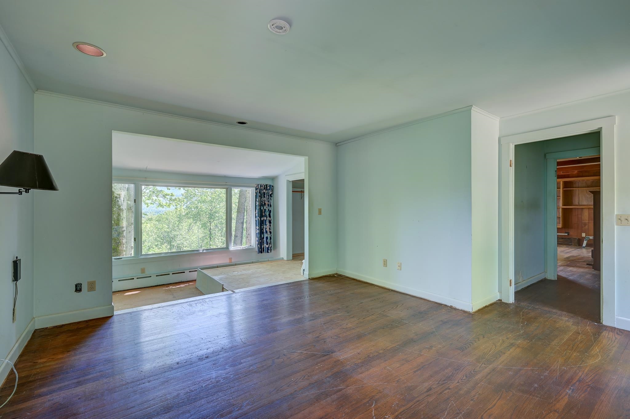 Vacant living area with worn hardwood floors, pale green walls, and a wide opening to a bright windowed room.