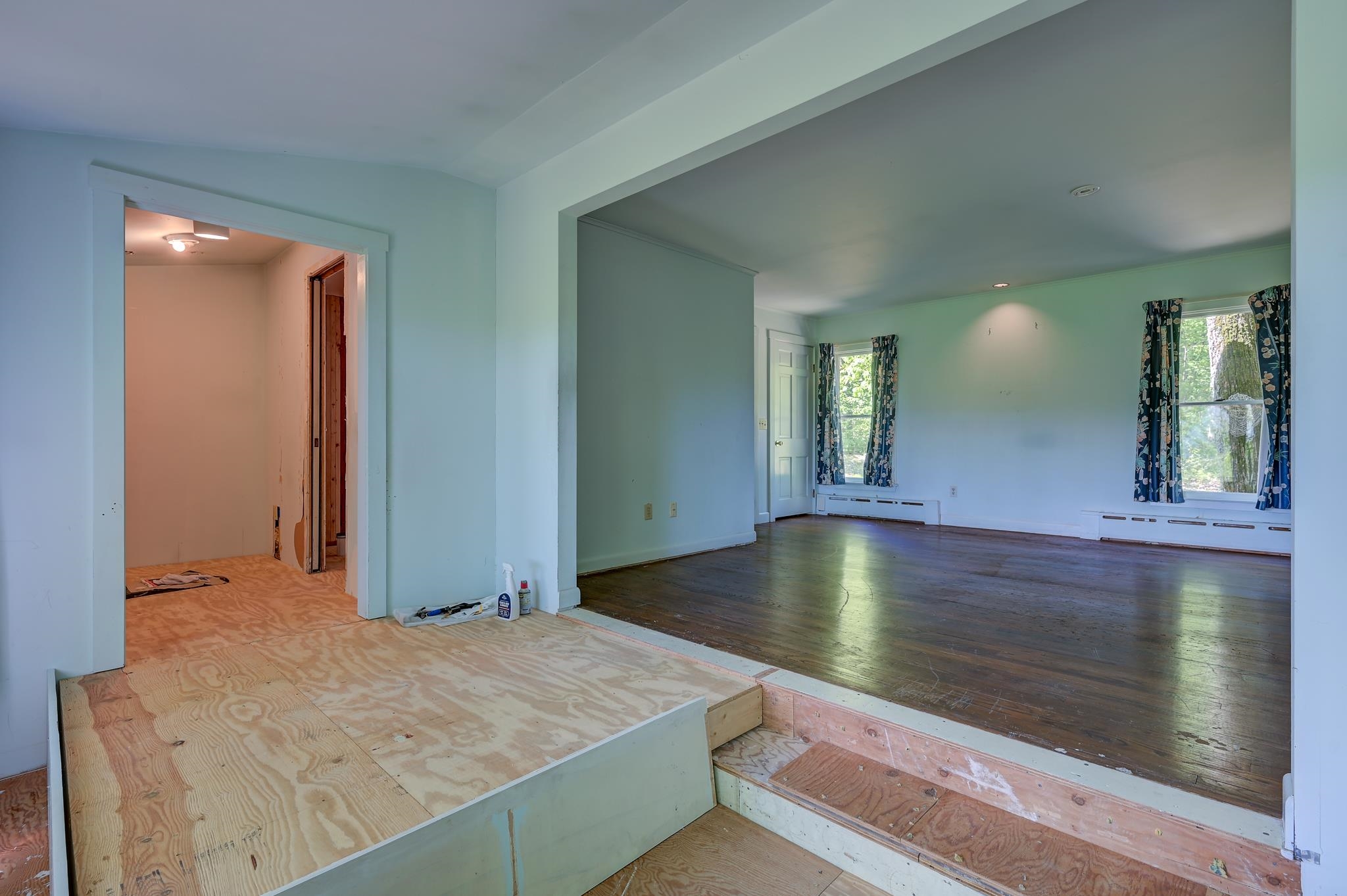 Vacant living room with dark wood floors, curtained windows, and exposed plywood flooring under renovation.