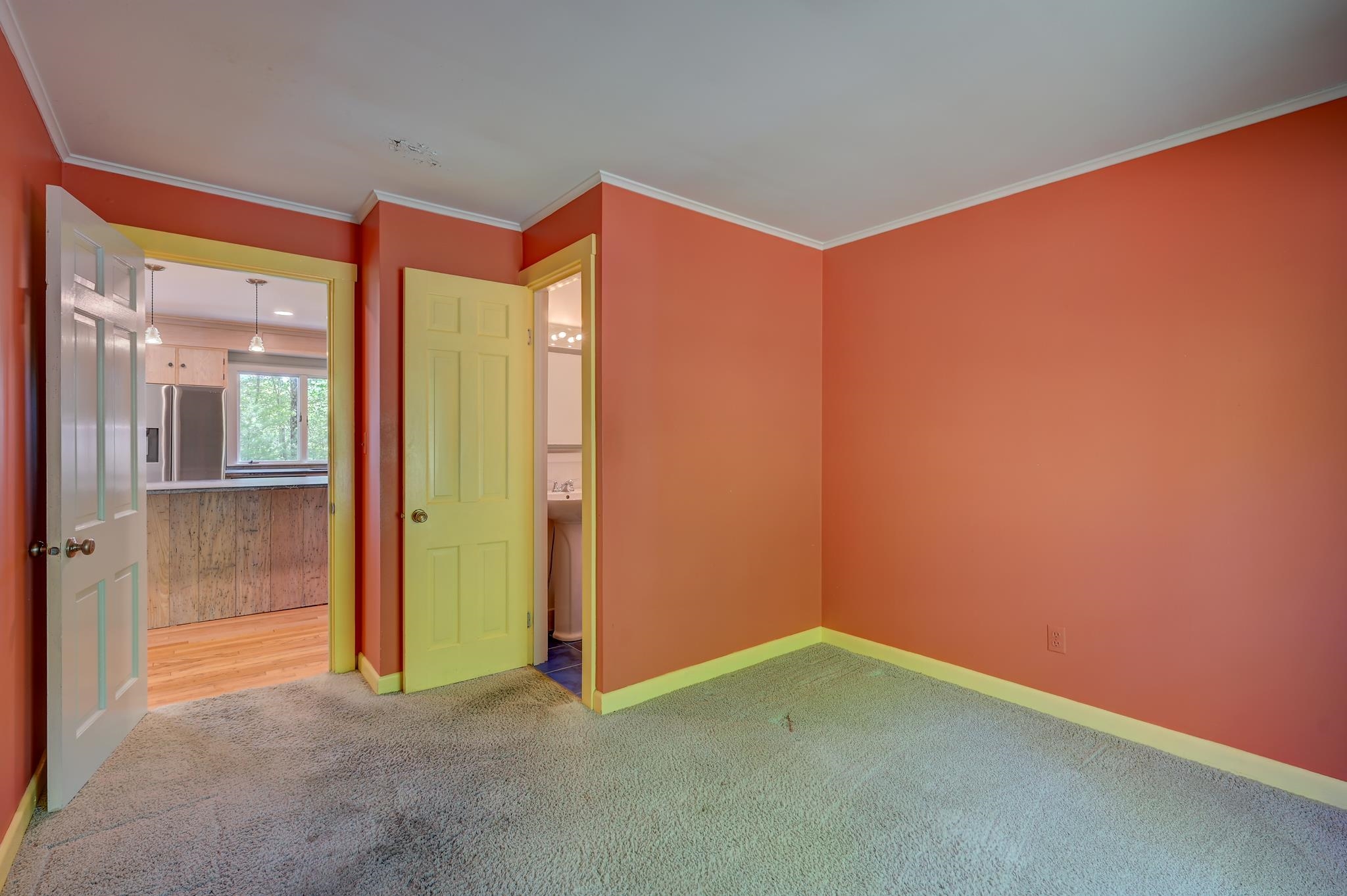 Empty bedroom with coral walls, yellow-green trim, beige carpet, and open doors to the kitchen and bathroom.