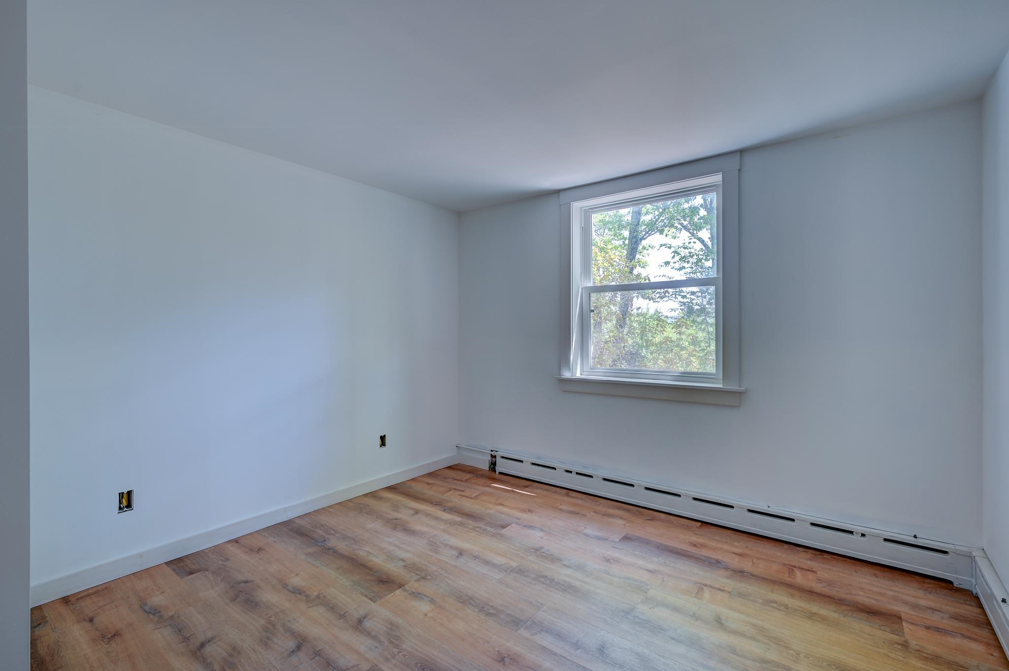 Empty bedroom with wood-look flooring, a window overlooking trees, and a long baseboard heater.