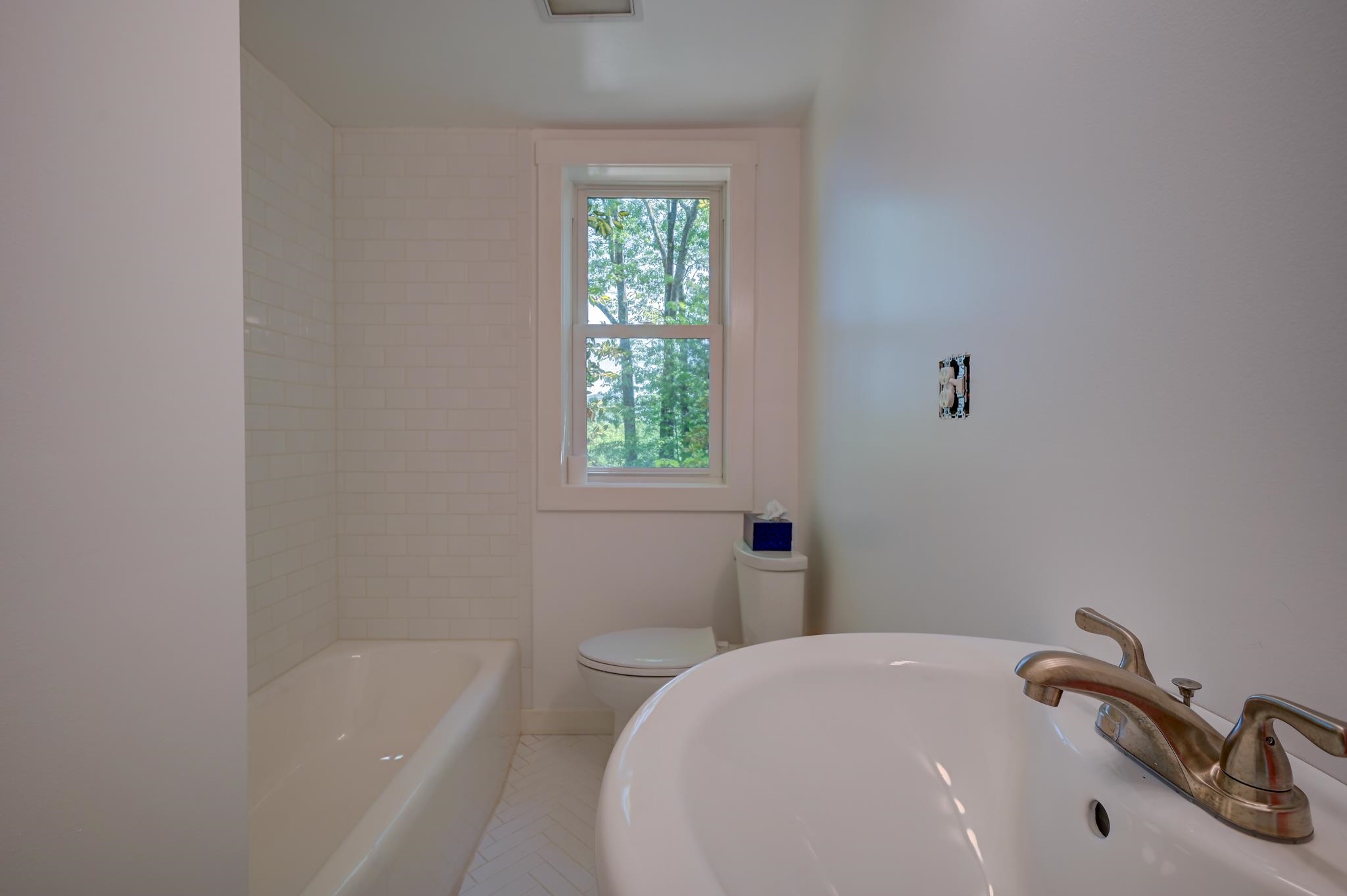 Bathroom with tiled bathtub, toilet, and sink beneath a window overlooking leafy trees.