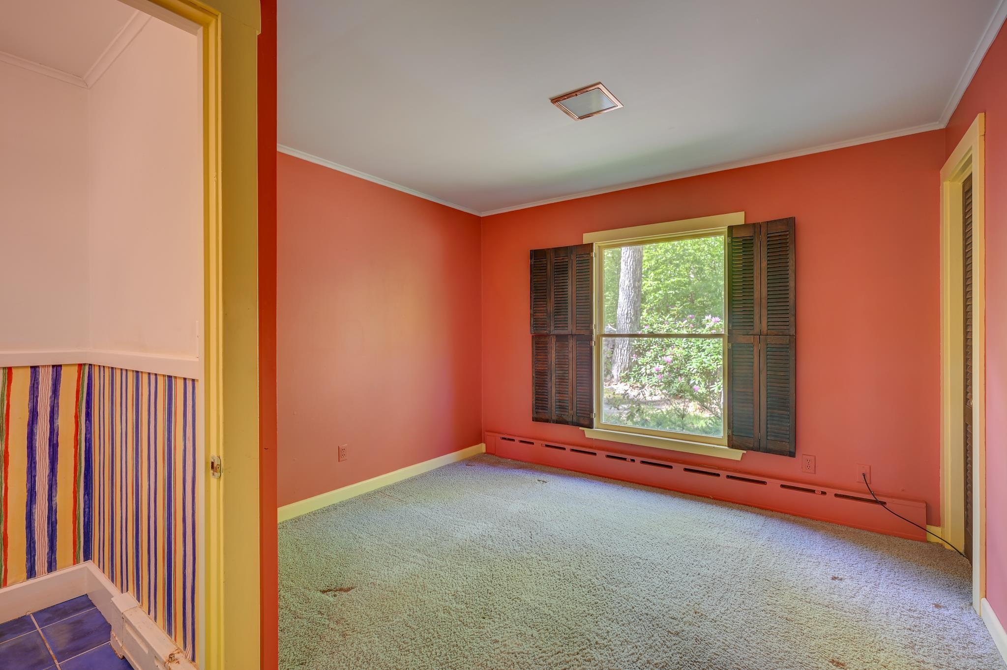 Empty coral-walled bedroom with carpet and shuttered window overlooking greenery.