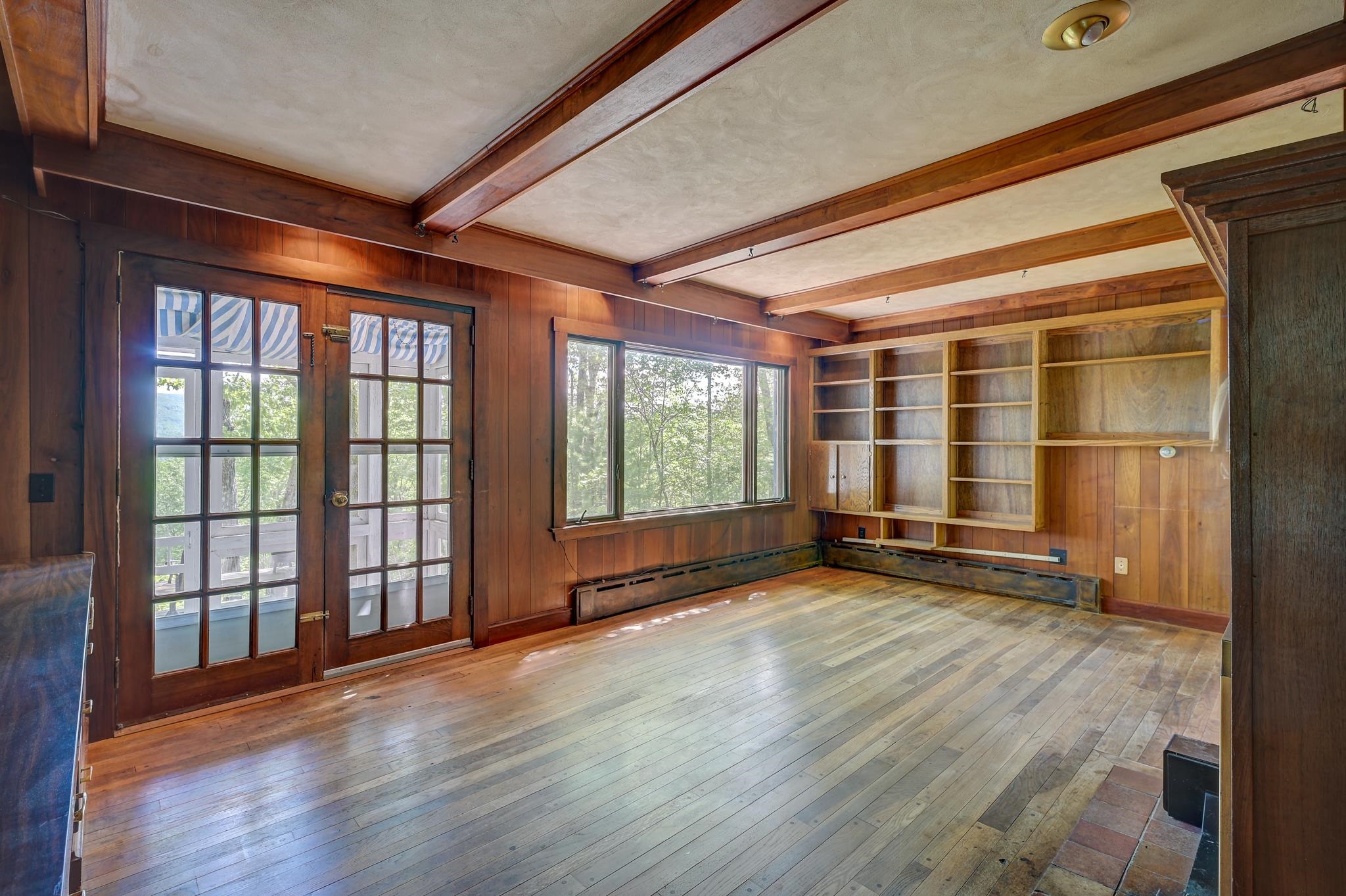Empty wood-paneled room with exposed beams, built-in shelves, hardwood floors, and glass-paneled doors.