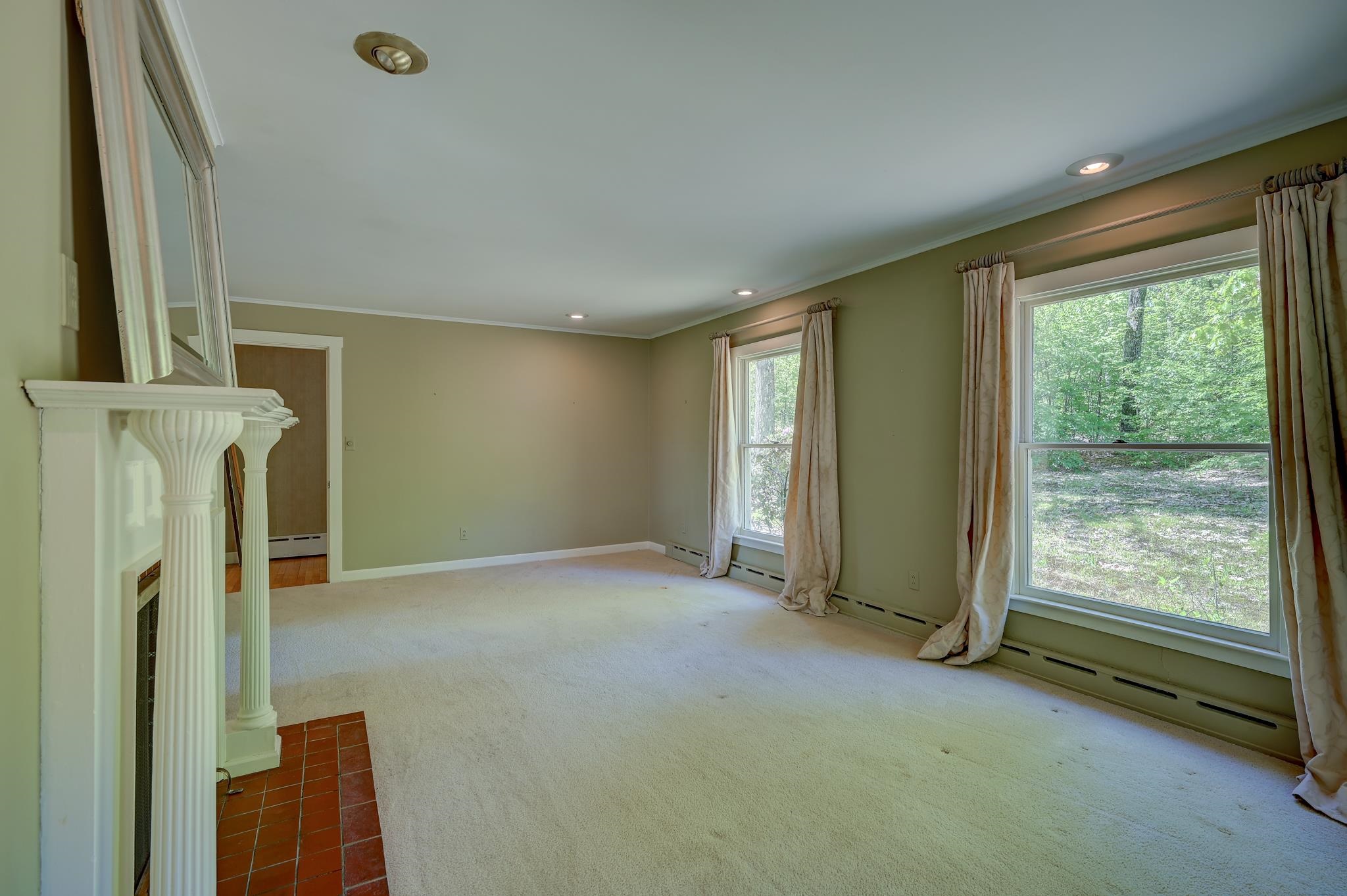 Empty living room with a white fireplace and two large windows overlooking trees.