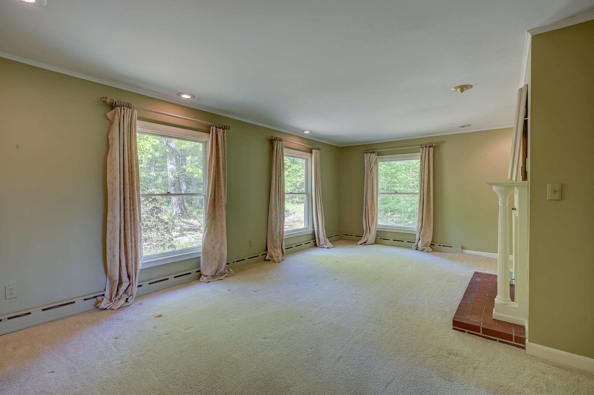 Empty living room with three curtained windows, pale carpet, and a brick fireplace hearth.