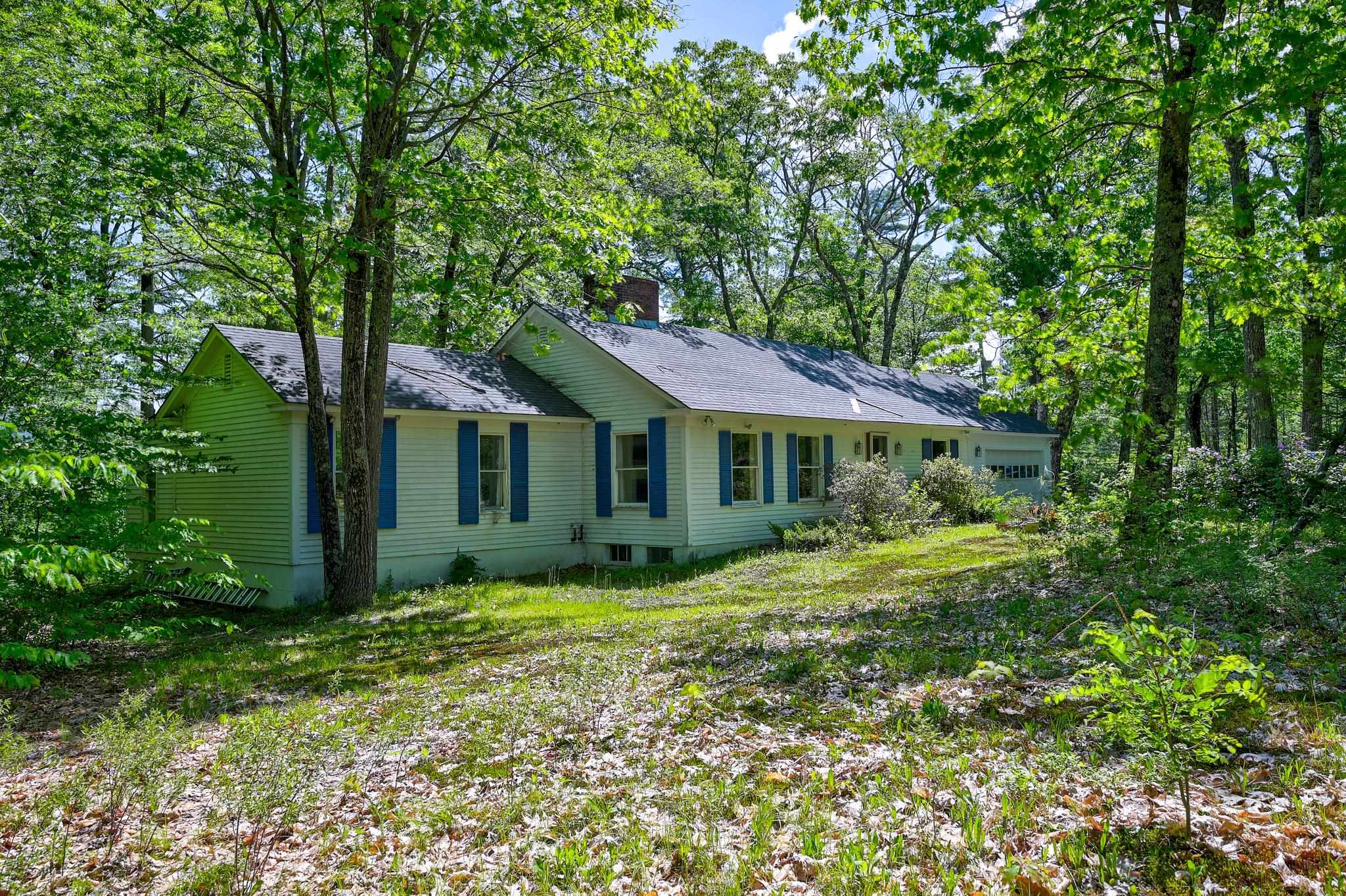 Light-colored house with blue shutters and an attached garage among mature trees.