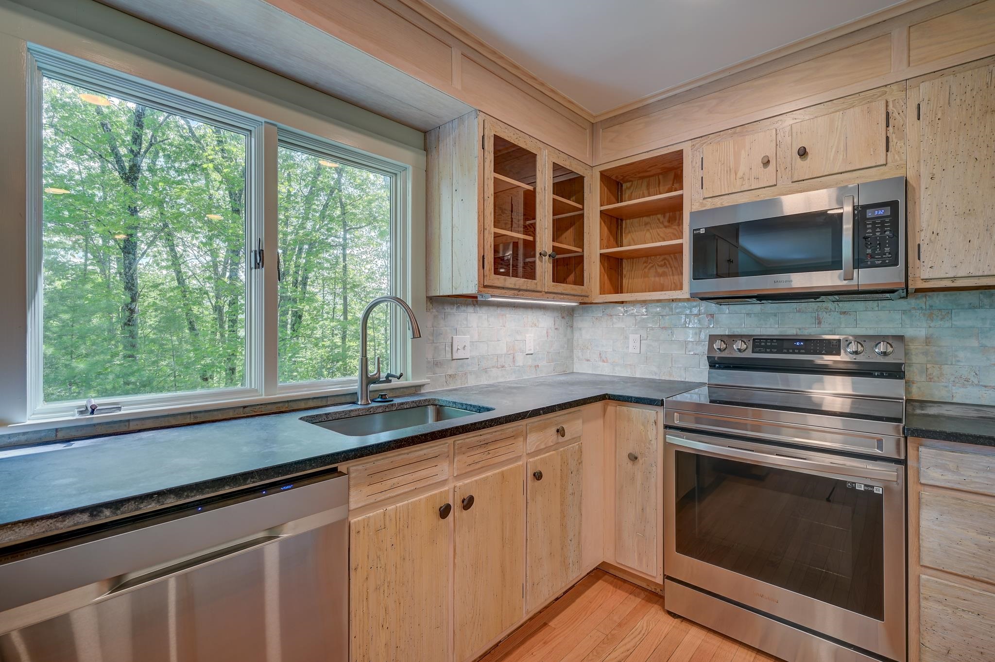 Kitchen with light wood cabinets, stainless steel appliances, and a sink beneath windows overlooking trees.