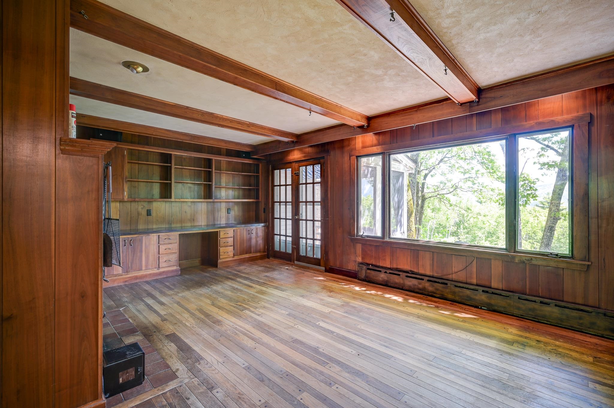 Empty wood-paneled room with built-in shelving, exposed beams, and large windows overlooking trees.