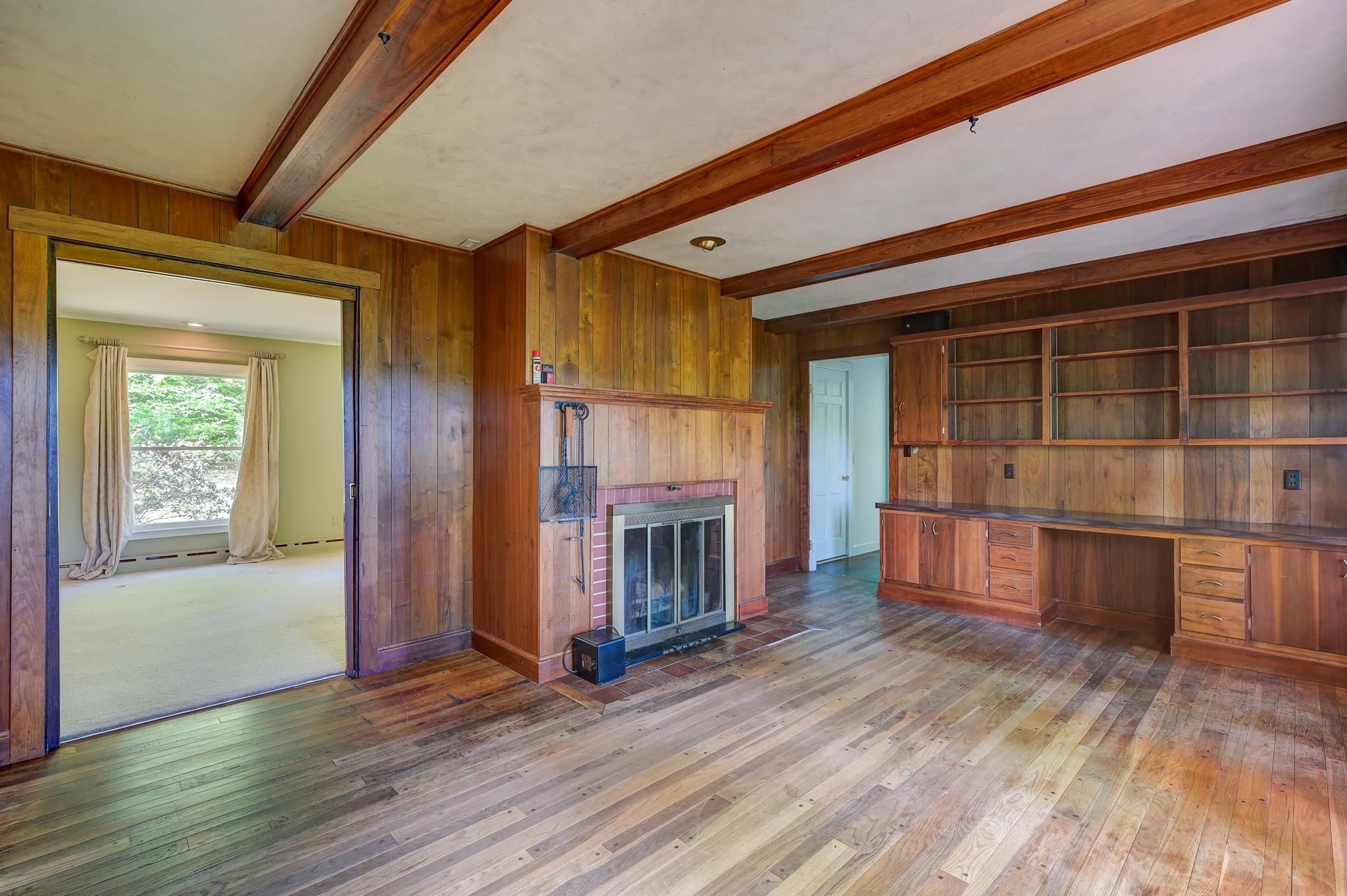 Wood-paneled living room with exposed beams, a brick fireplace, built-in shelving, and hardwood floors.