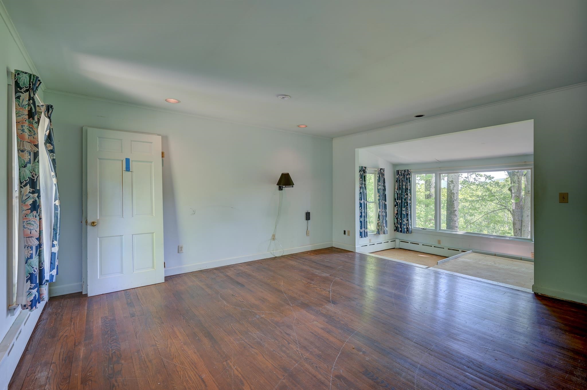 Empty living room with worn hardwood floors and large windows overlooking trees.