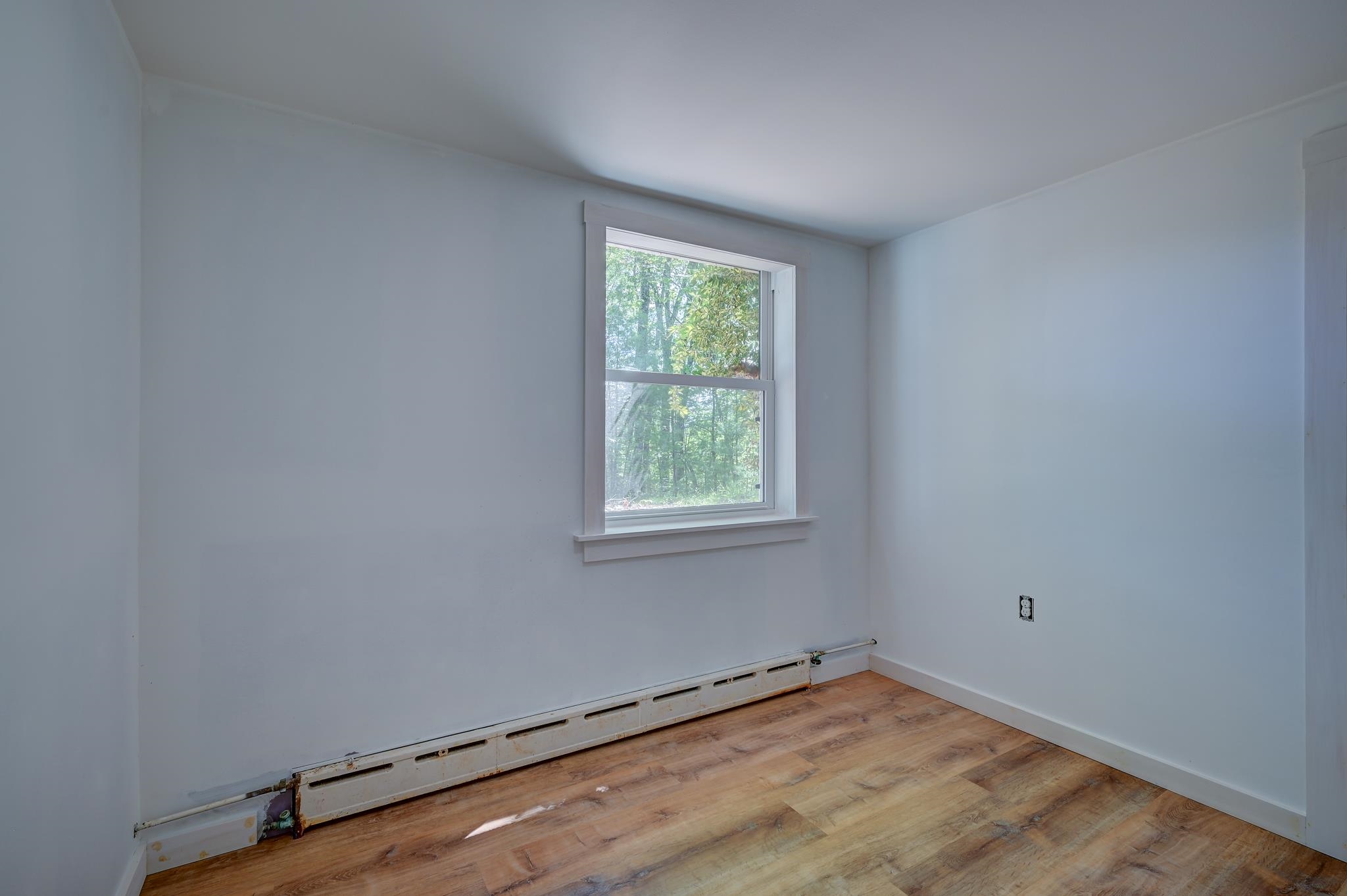 Empty room with wood-look flooring, a window overlooking trees, and exposed baseboard heating.