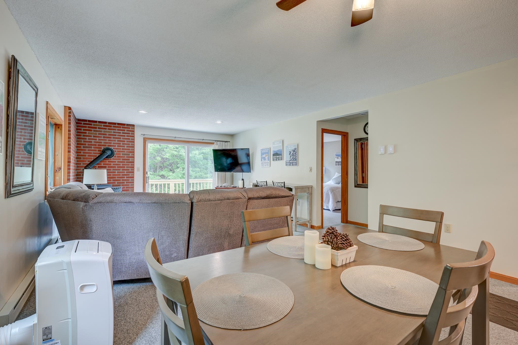 Open living and dining area with a brick wall, gray sofa, and sliding doors to a leafy view.