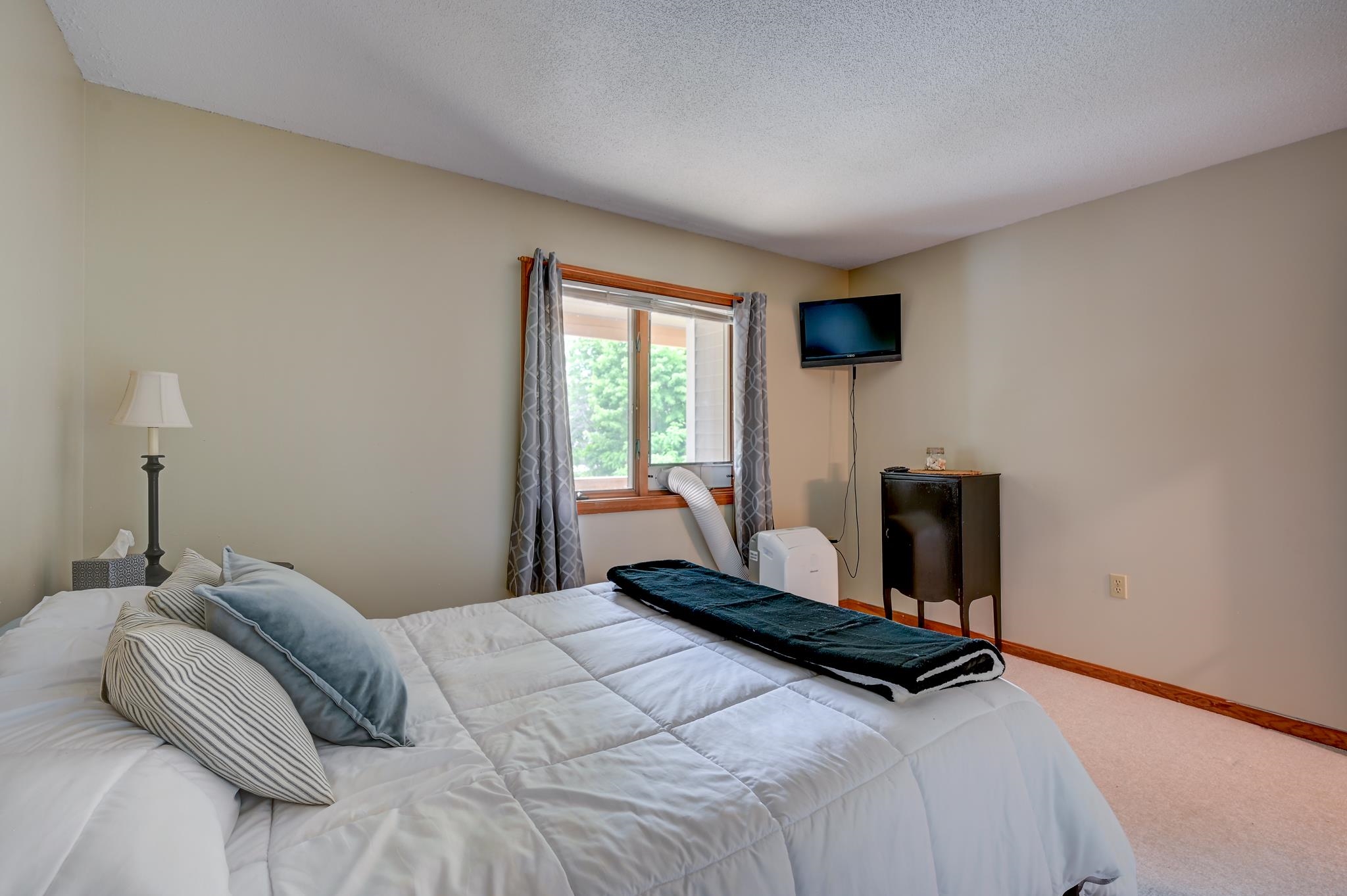 Bedroom with a white bed, gray pillows, curtained window, portable air conditioner, and wall-mounted television.