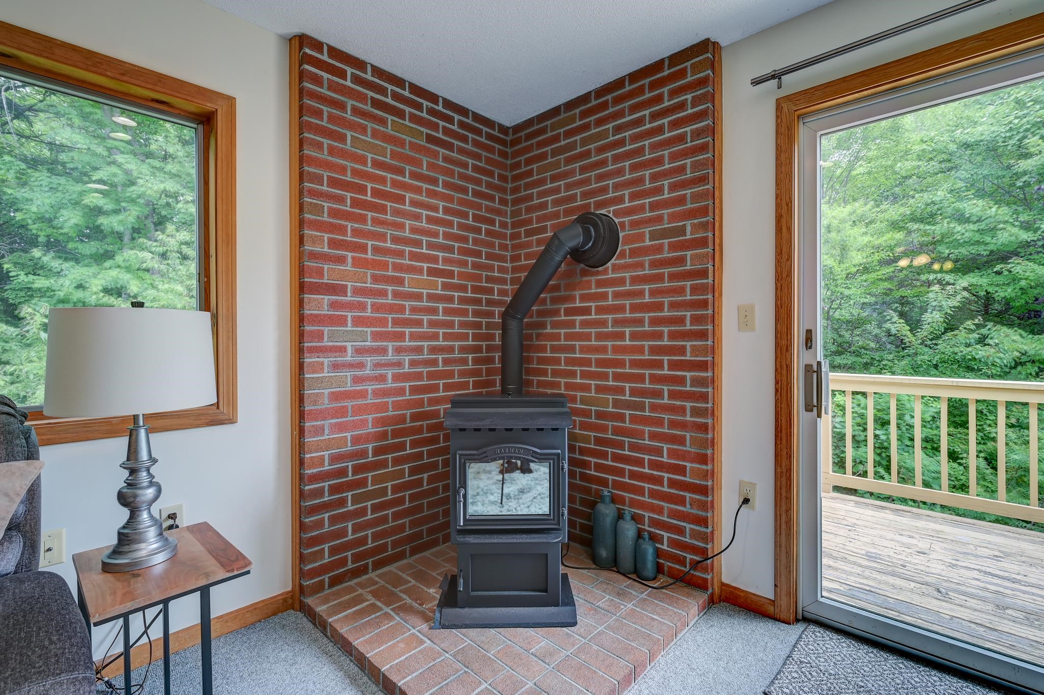 Freestanding wood stove against a brick hearth, beside a sliding door opening onto a wooded deck.