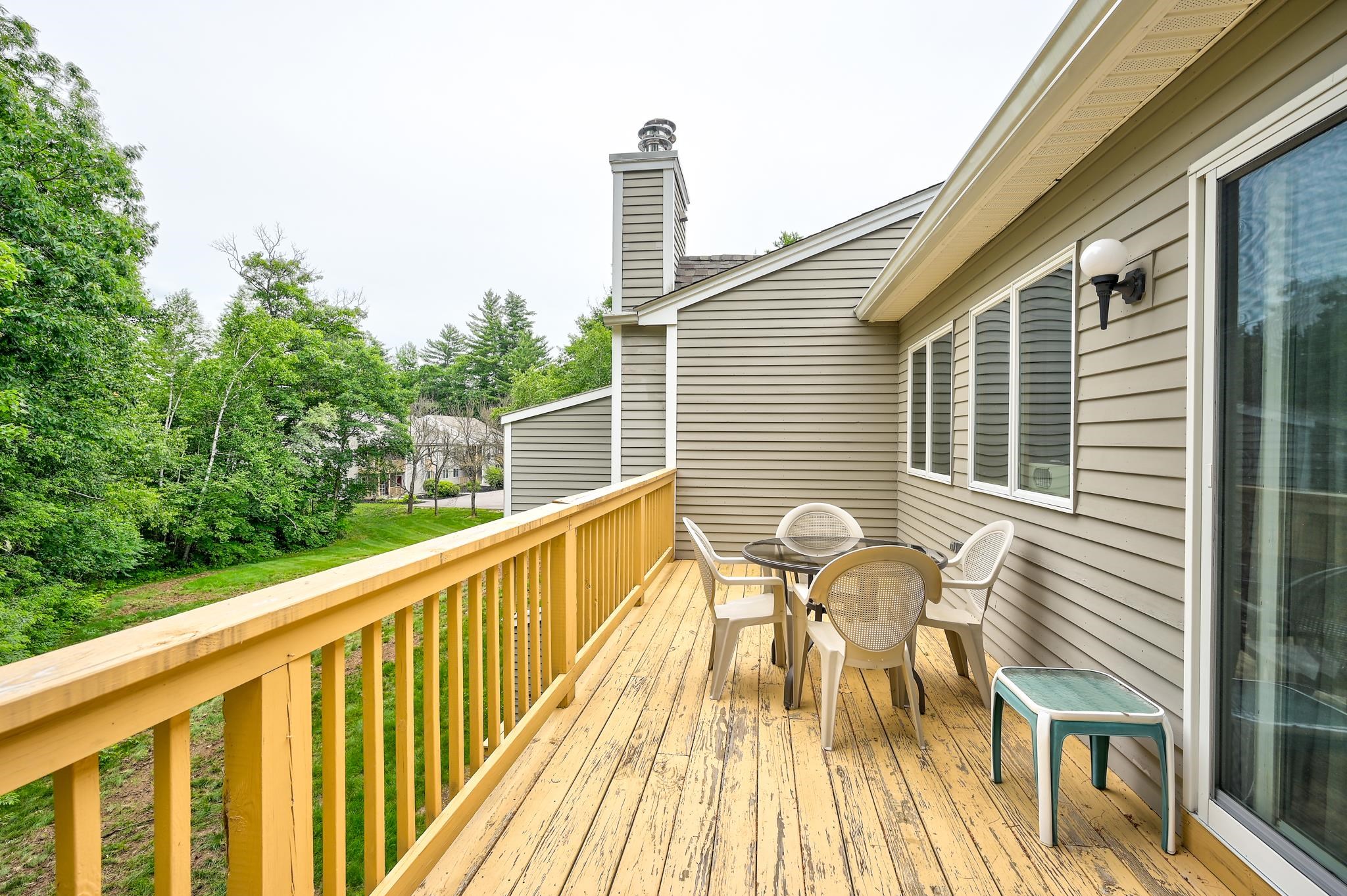 Wood deck with outdoor dining set and yellow railing beside a wooded lawn.