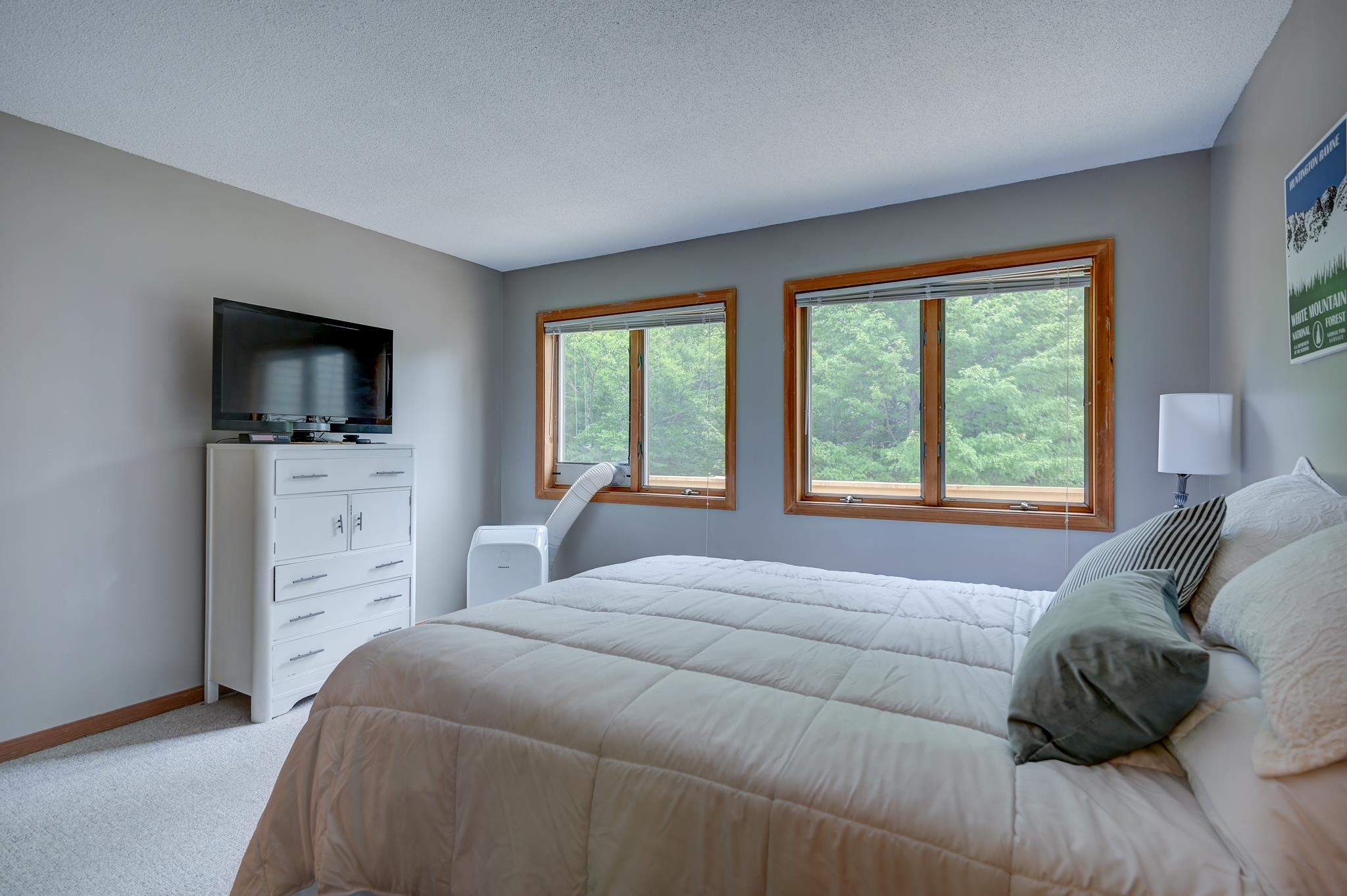 Bedroom with a quilted bed, white dresser and television, and windows overlooking trees.