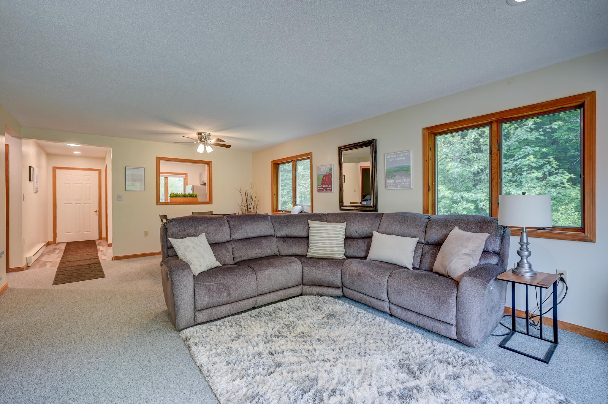 Furnished living room with a gray sectional, pale rug, and large windows facing leafy trees.
