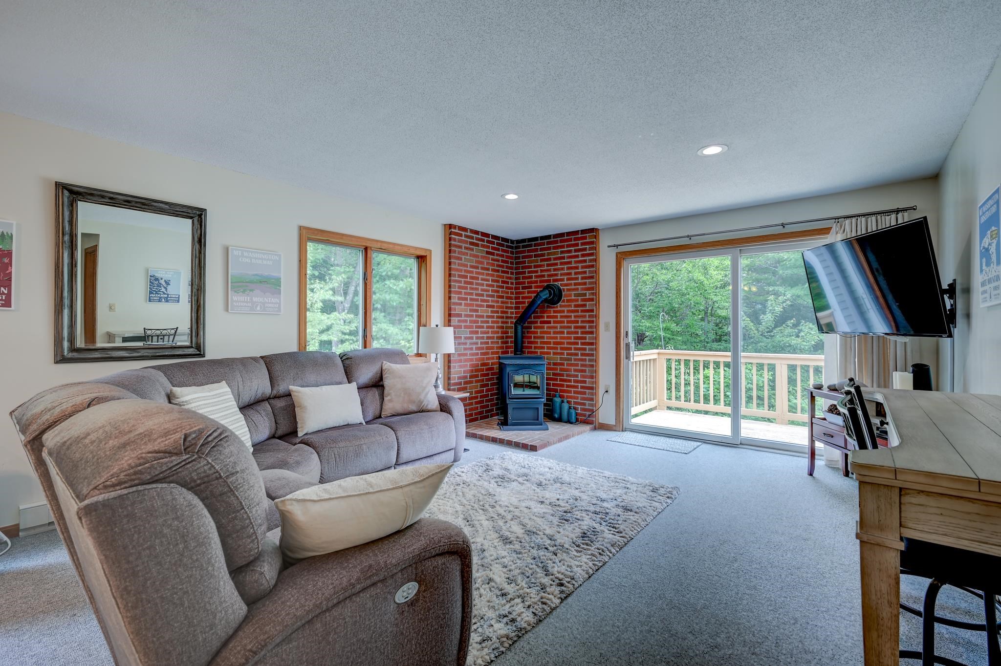 Living room with a gray sectional, brick-corner wood stove, and sliding doors to a wooded deck.