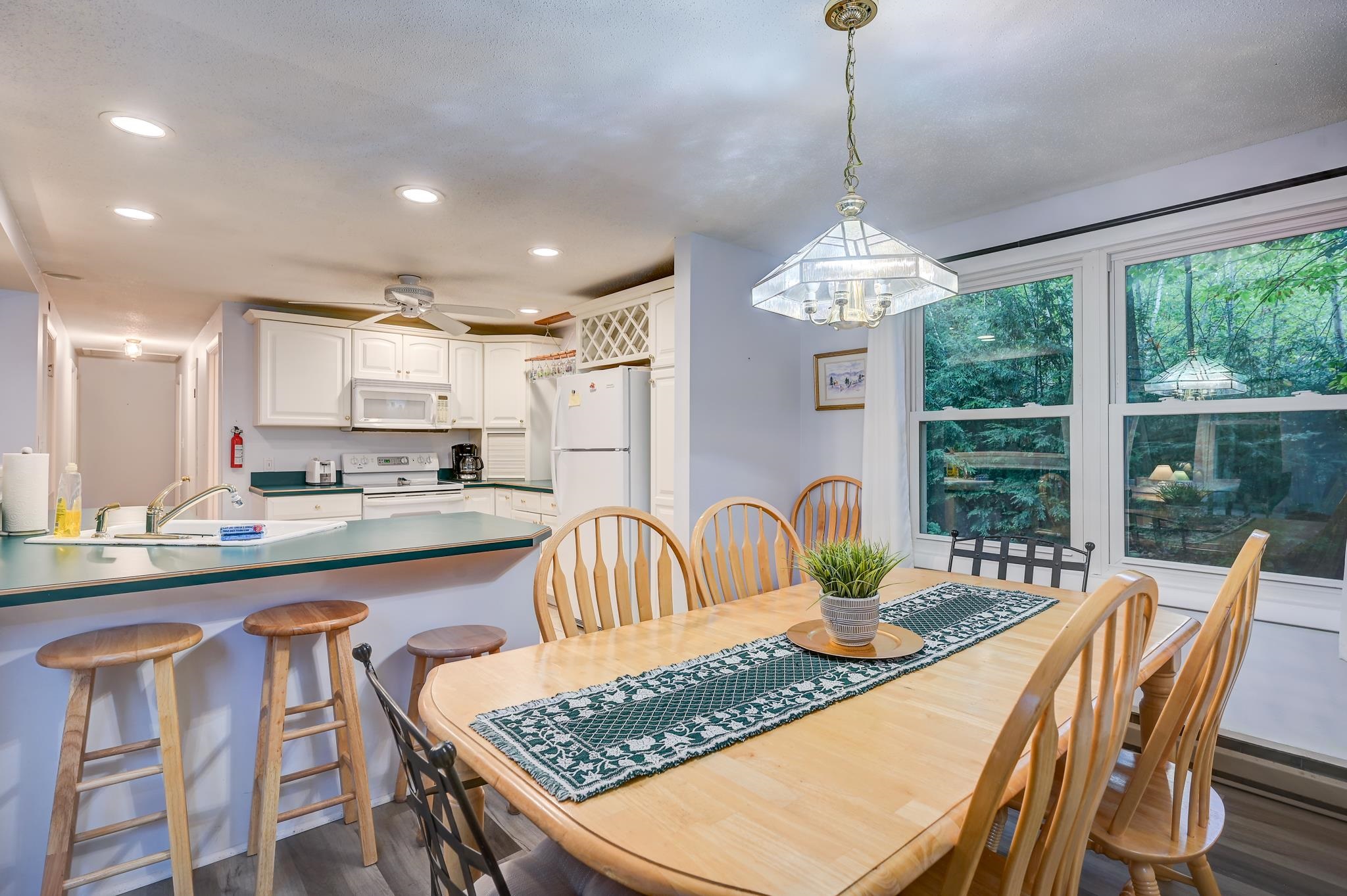 Kitchen and dining area with a breakfast bar, wooden table, and large windows overlooking trees.
