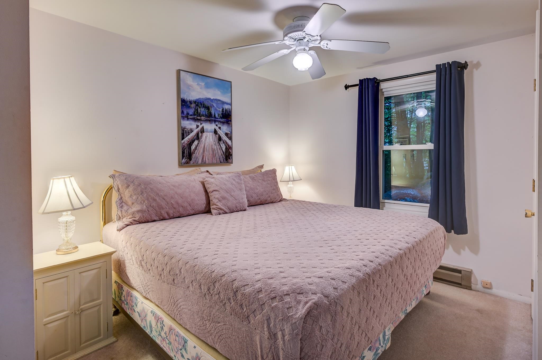 Furnished bedroom with a mauve quilted bed, dark window curtains, and a ceiling fan.