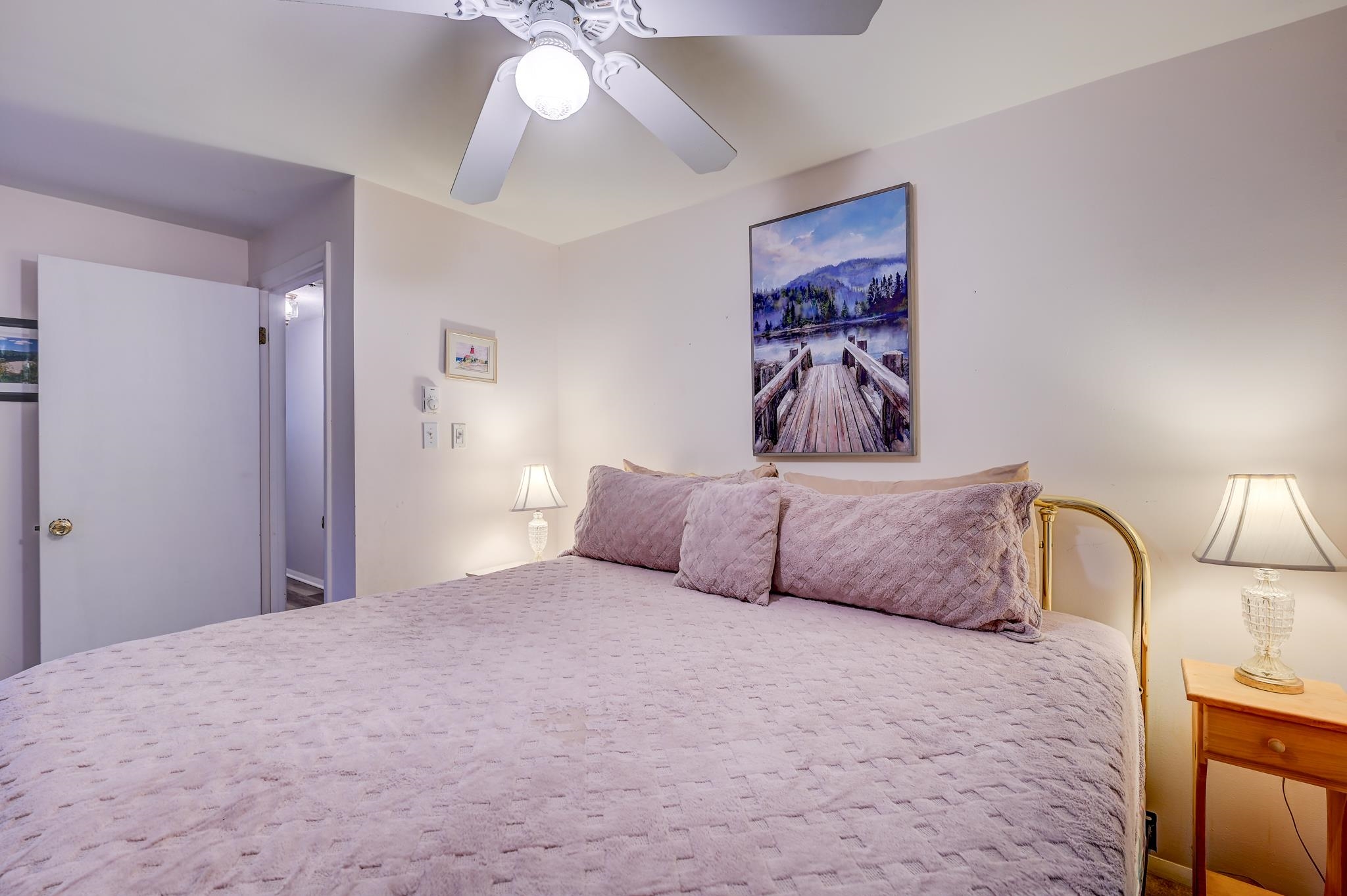Bedroom with a lavender-covered bed, brass headboard, matching lamps, and framed lakeside artwork.