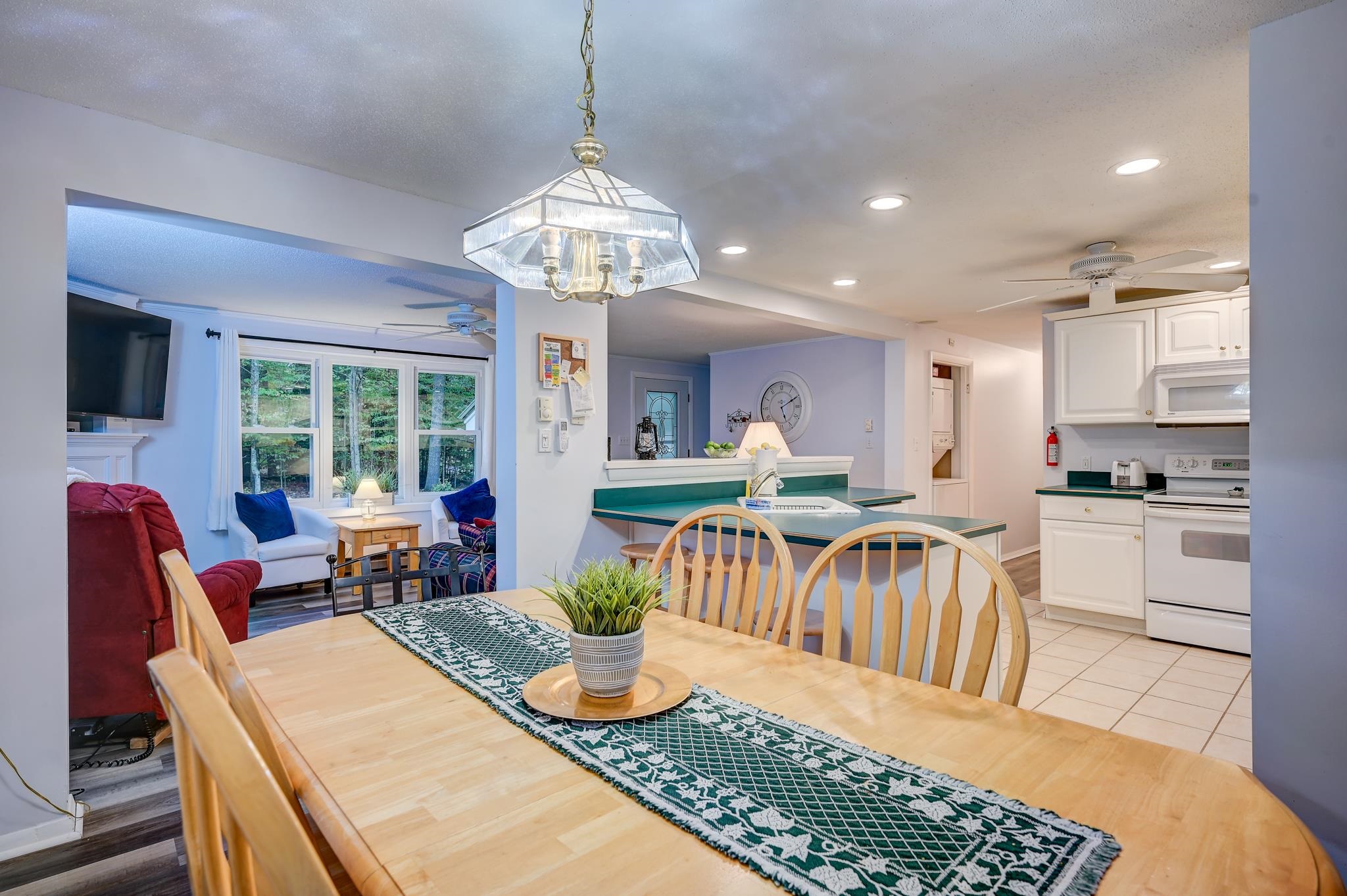 Open-plan kitchen and dining area with a teal peninsula, wooden table, and windowed sitting area.