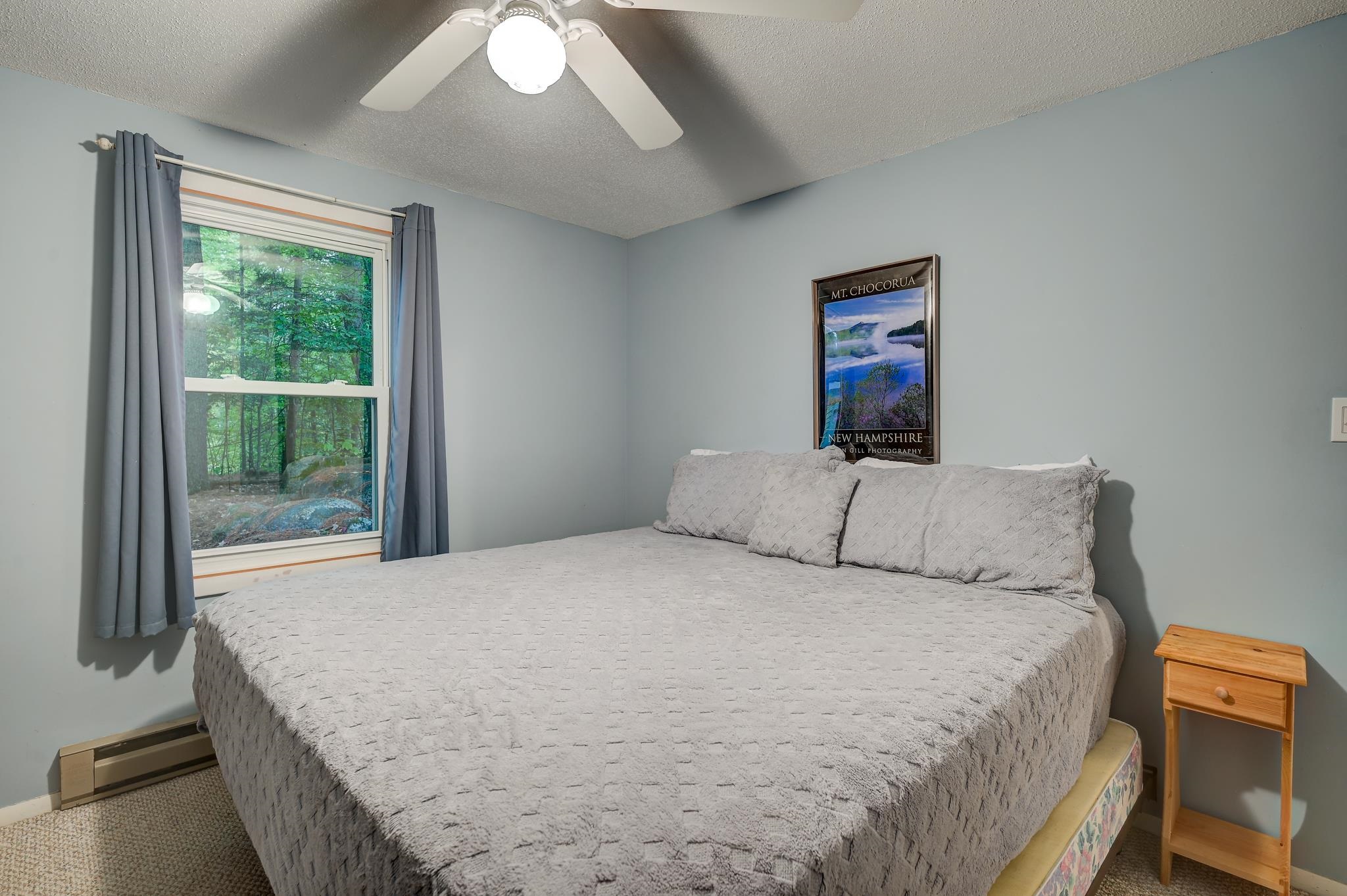 Bedroom with a large gray-covered bed, wooded window view, and Mount Chocorua poster.