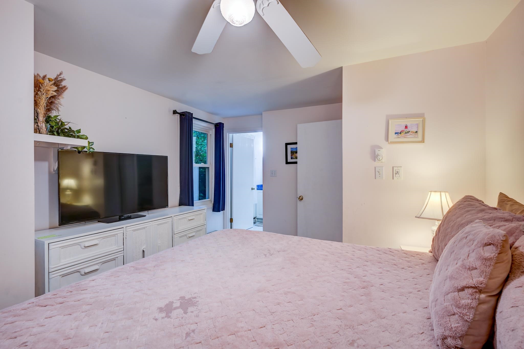 Furnished bedroom with a large pink-covered bed, television on a white dresser, and navy curtains.