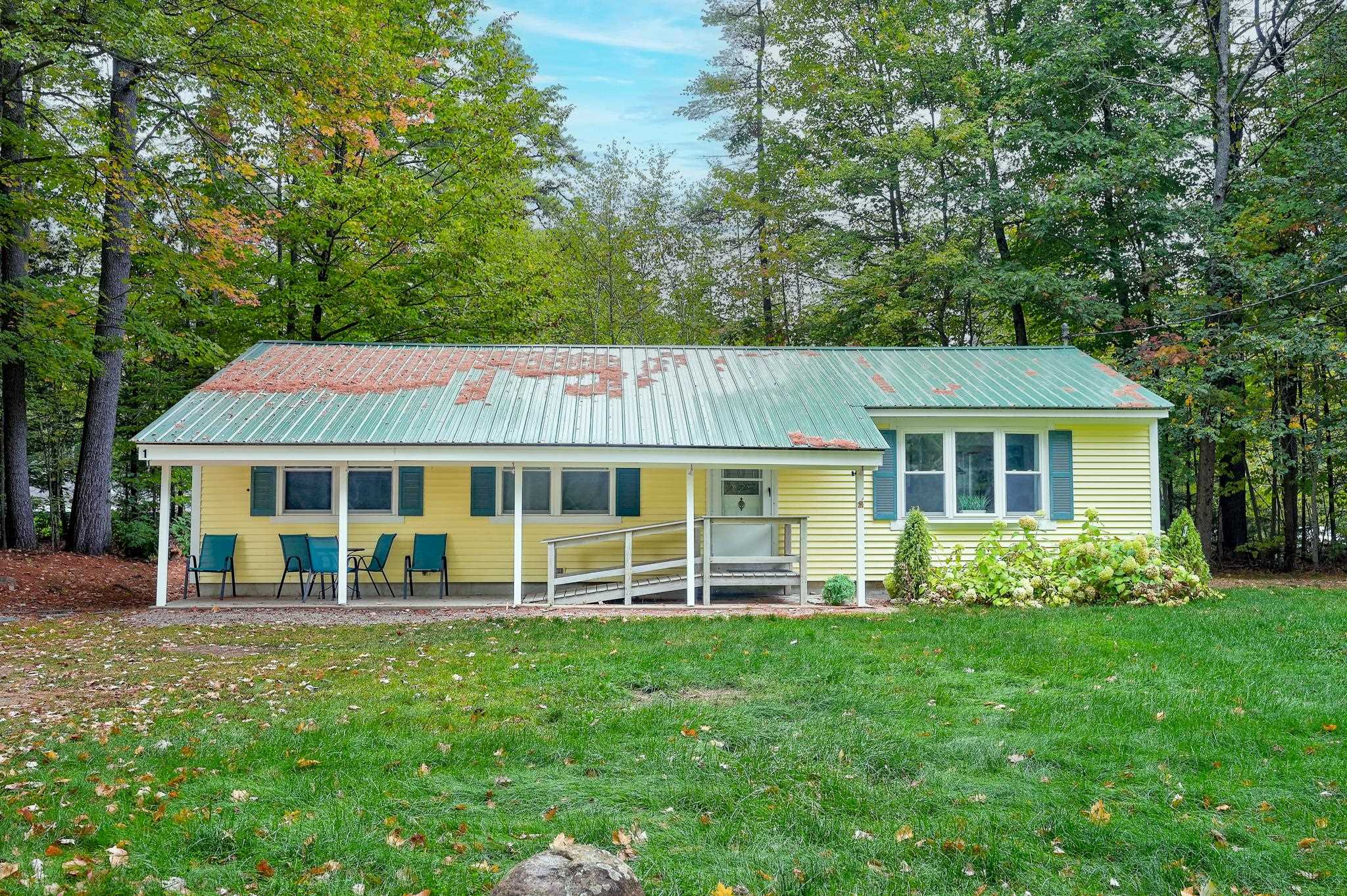 Yellow single-story home with a covered porch, accessibility ramp, and weathered metal roof among trees.