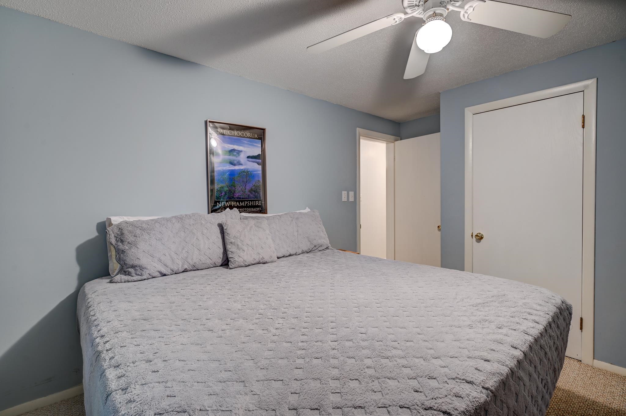 Bedroom with a large gray-covered bed, blue-gray walls, ceiling fan, and framed Mt. Chocorua poster.
