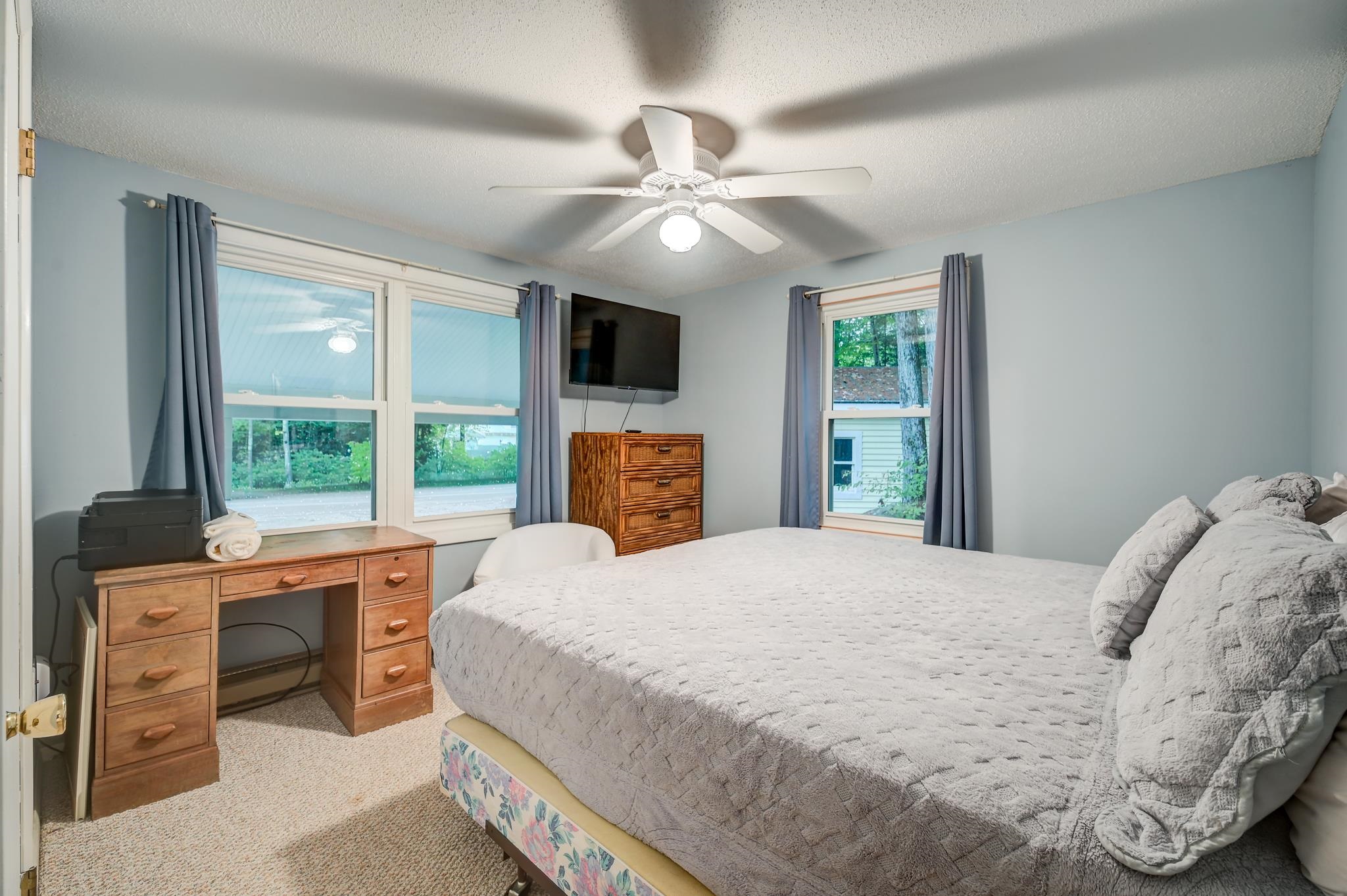 Bedroom with a large gray-covered bed, two windows, wooden furniture, and a ceiling fan.