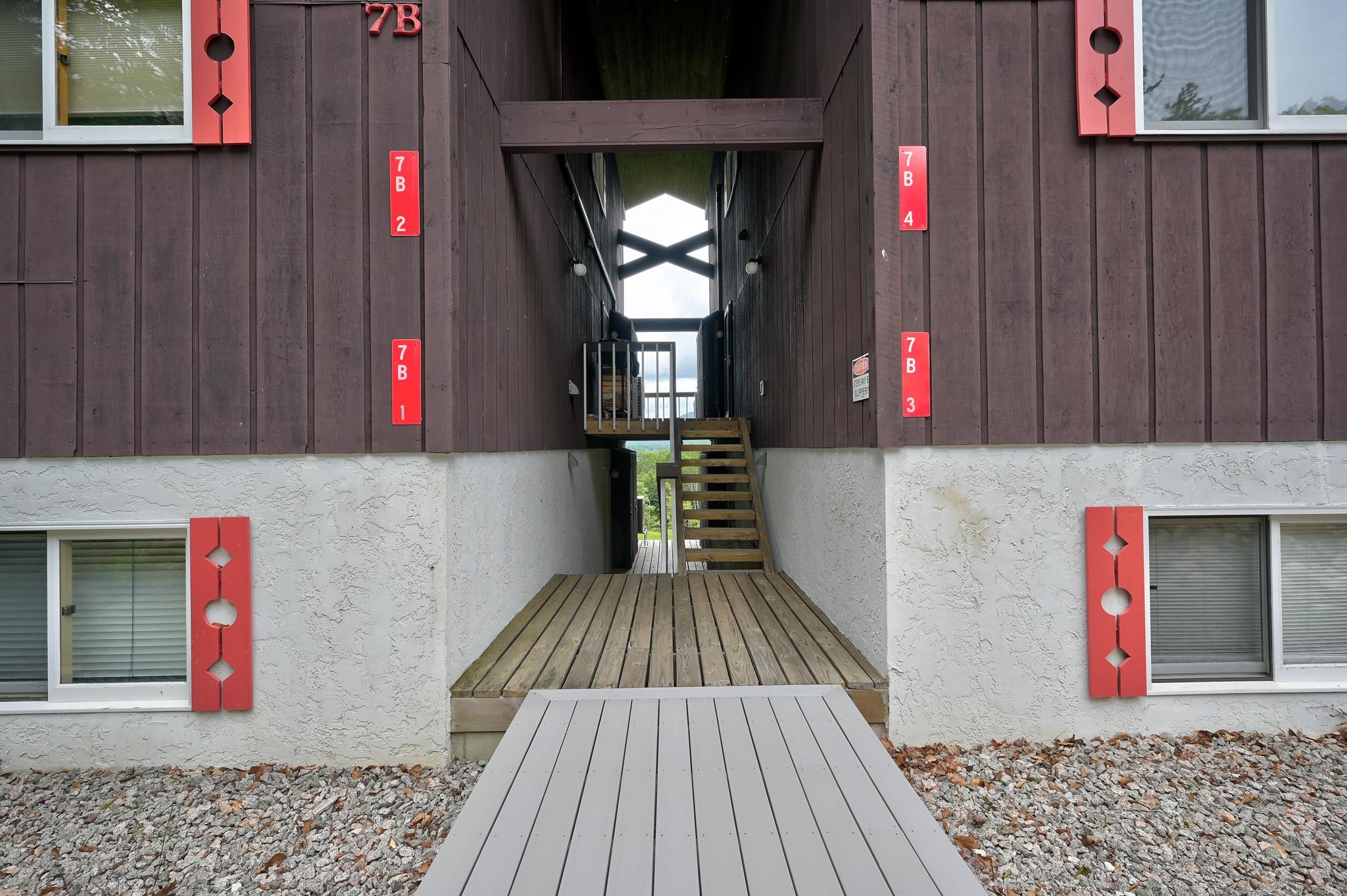 Building entrance with a central wooden walkway and stairs, dark siding, and red unit-number plaques.