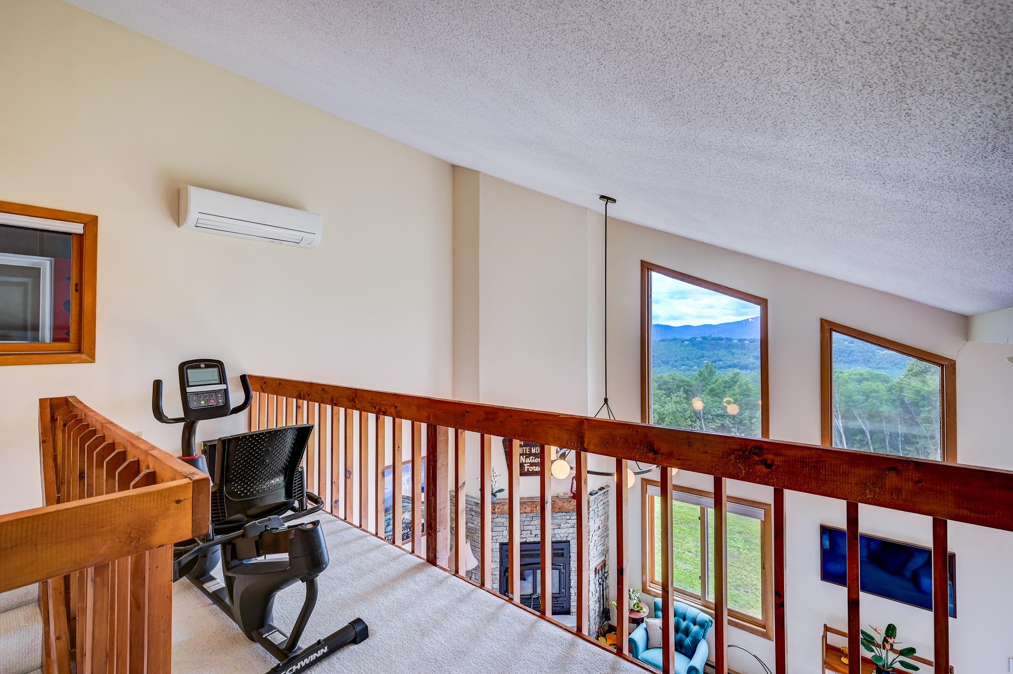 Furnished loft with an exercise bike, wooden railing, and mountain views through tall windows.