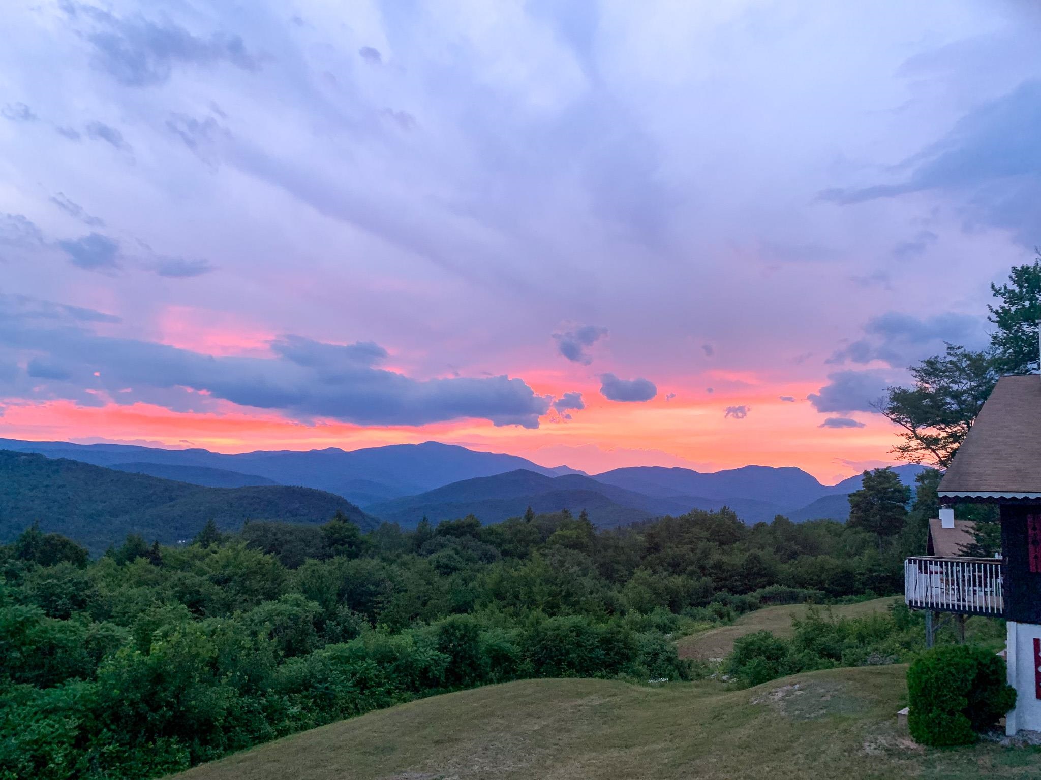 Sunset over layered mountains beside a home’s deck and grassy hillside.