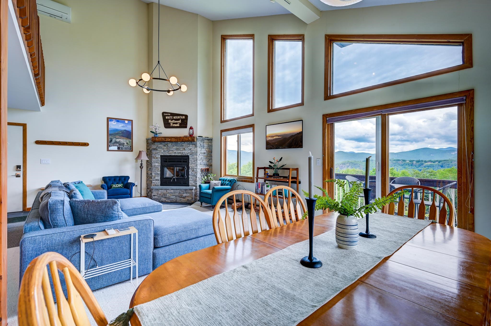 Open-plan living and dining room with a stone fireplace and mountain views through tall windows.