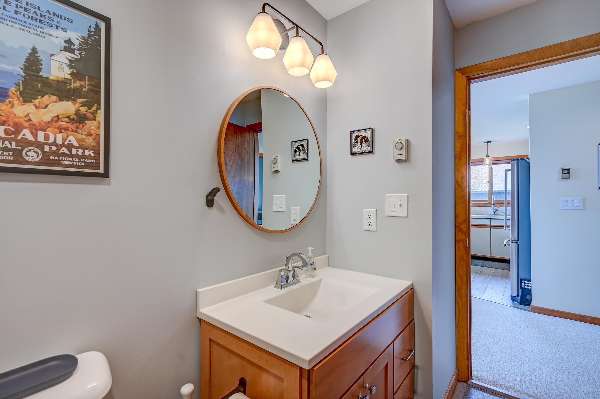 Bathroom vanity with a white sink, wood cabinetry, oval mirror, and doorway to the kitchen.