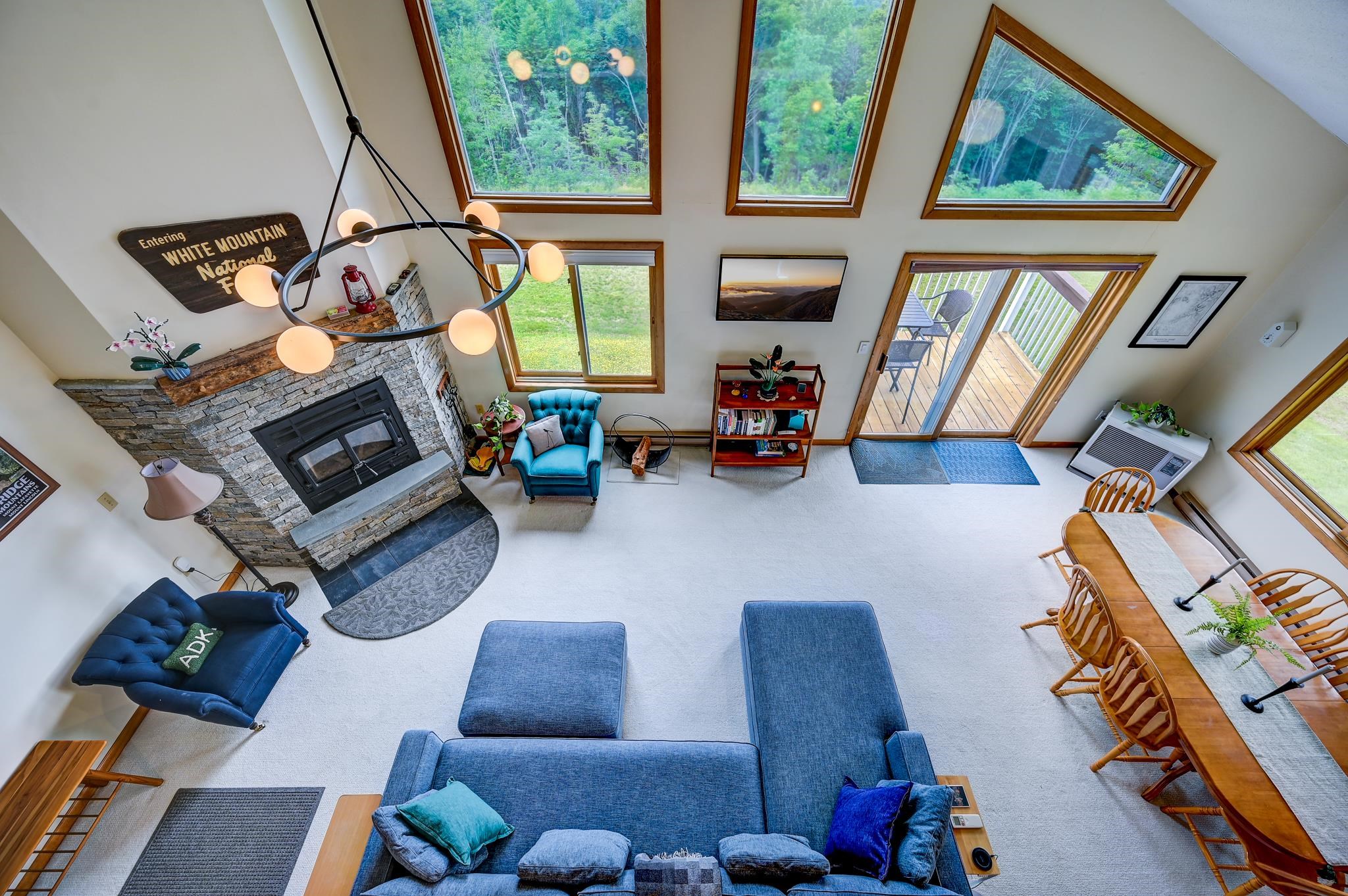 Vaulted living room with a stone fireplace, blue seating, and tall windows overlooking trees.