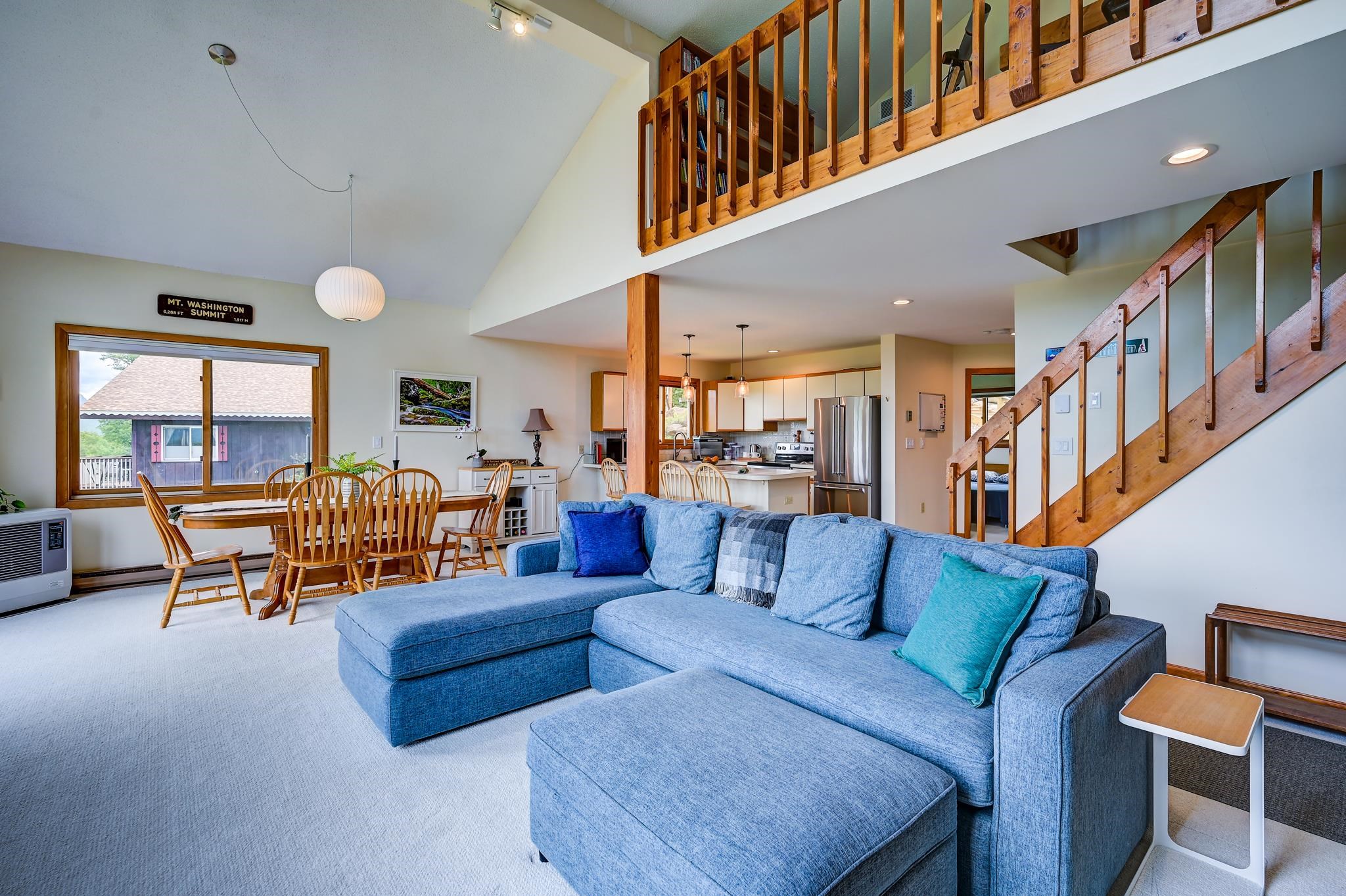 Open-plan living room with a large blue sectional, wooden staircase, loft railing, dining area, and kitchen.