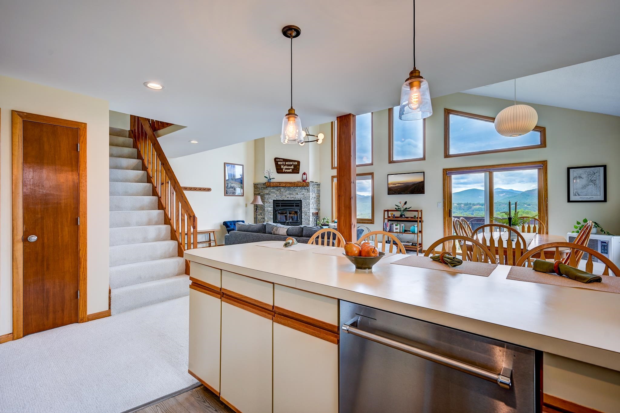 Open-plan kitchen and living room with a stone fireplace, dining table, and mountain views through tall windows.
