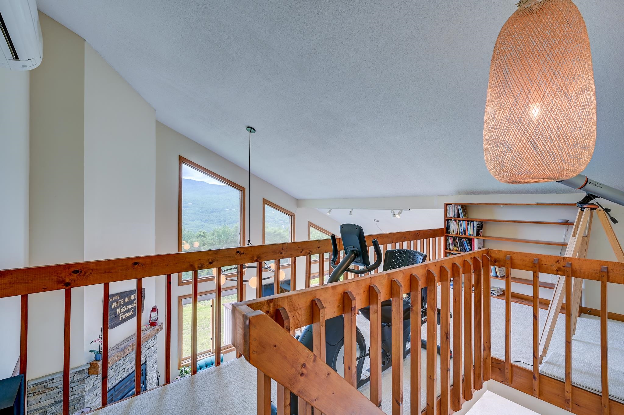 Loft with wood railings, exercise bike, telescope, bookshelves, and mountain views through tall windows.